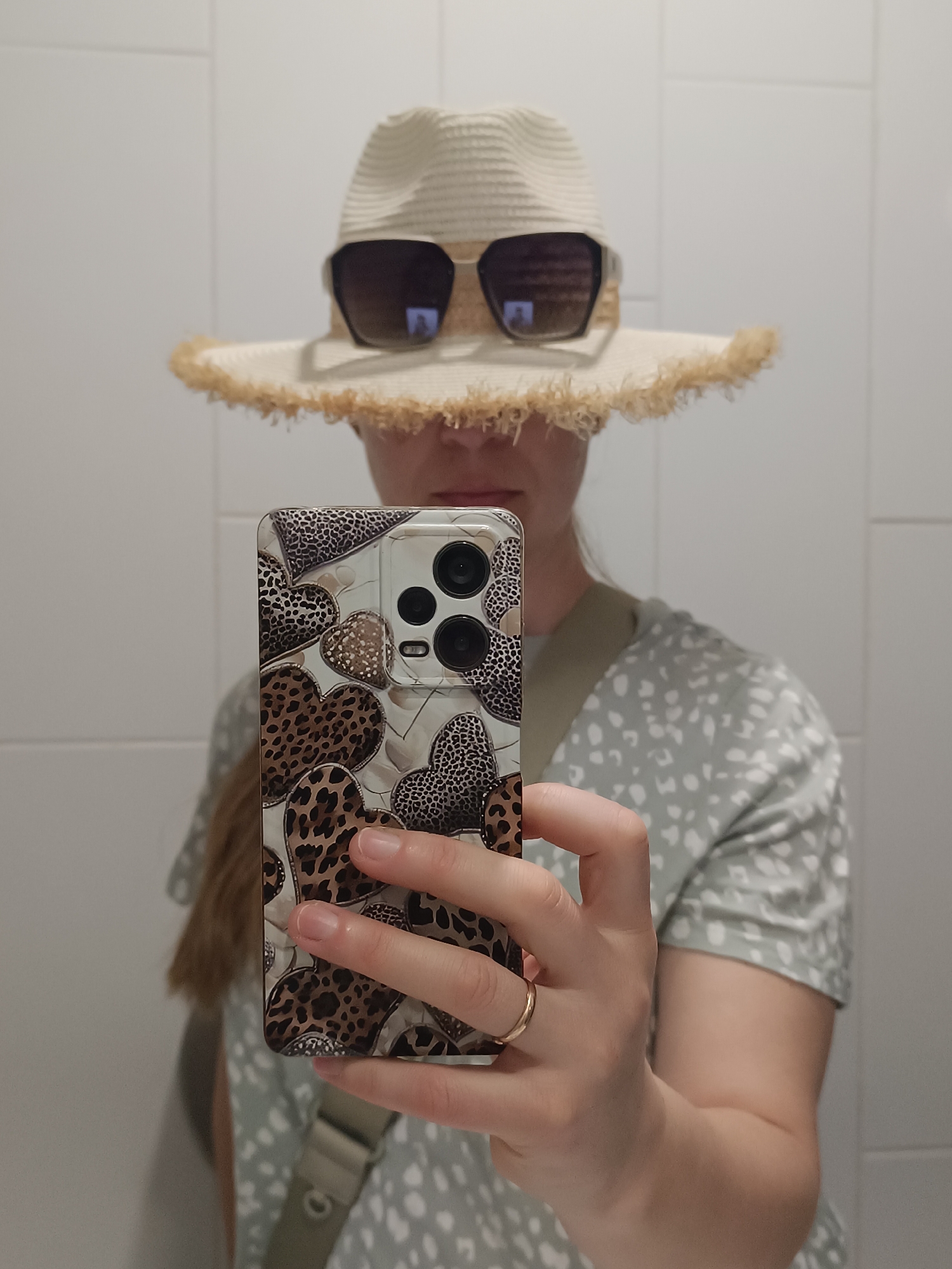 A person wearing a straw hat and sunglasses, partially obscuring their face, holds up a smartphone to take a mirror selfie. The phone case has a leopard print and heart design. The person is wearing a light green patterned t-shirt and a gold ring on their left hand. The background appears to be a tiled wall.