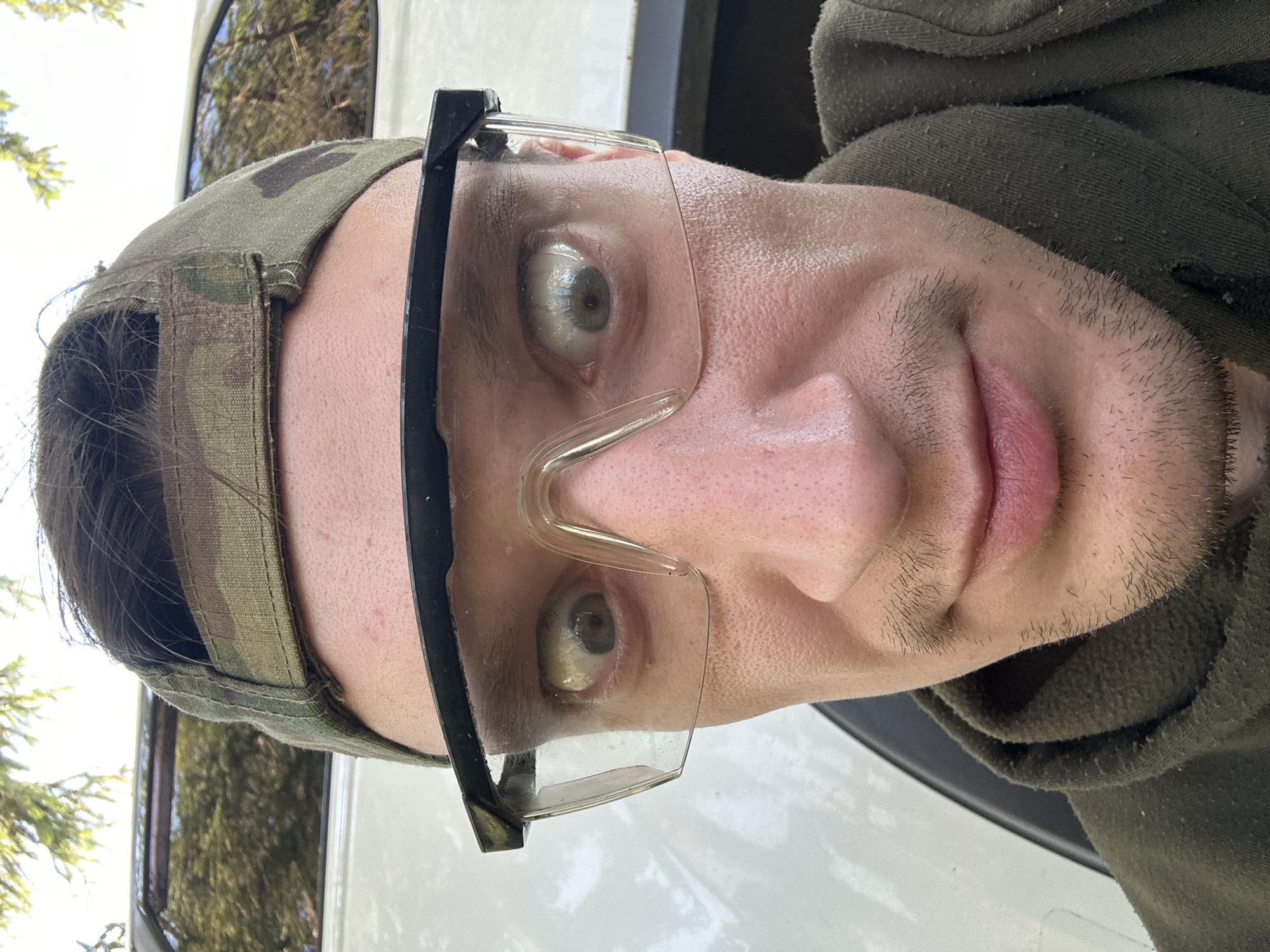 A close-up, eye-level, over-the-shoulder selfie shows a light-skinned person wearing a camouflage baseball cap and clear safety glasses. The person's eyes are wide and appear dilated. The person is also wearing a dark green hooded sweatshirt. The background shows foliage and the side of a white vehicle. The image does not provide specific details that would allow for a determination of its location in the city of Makiv, Ukraine.