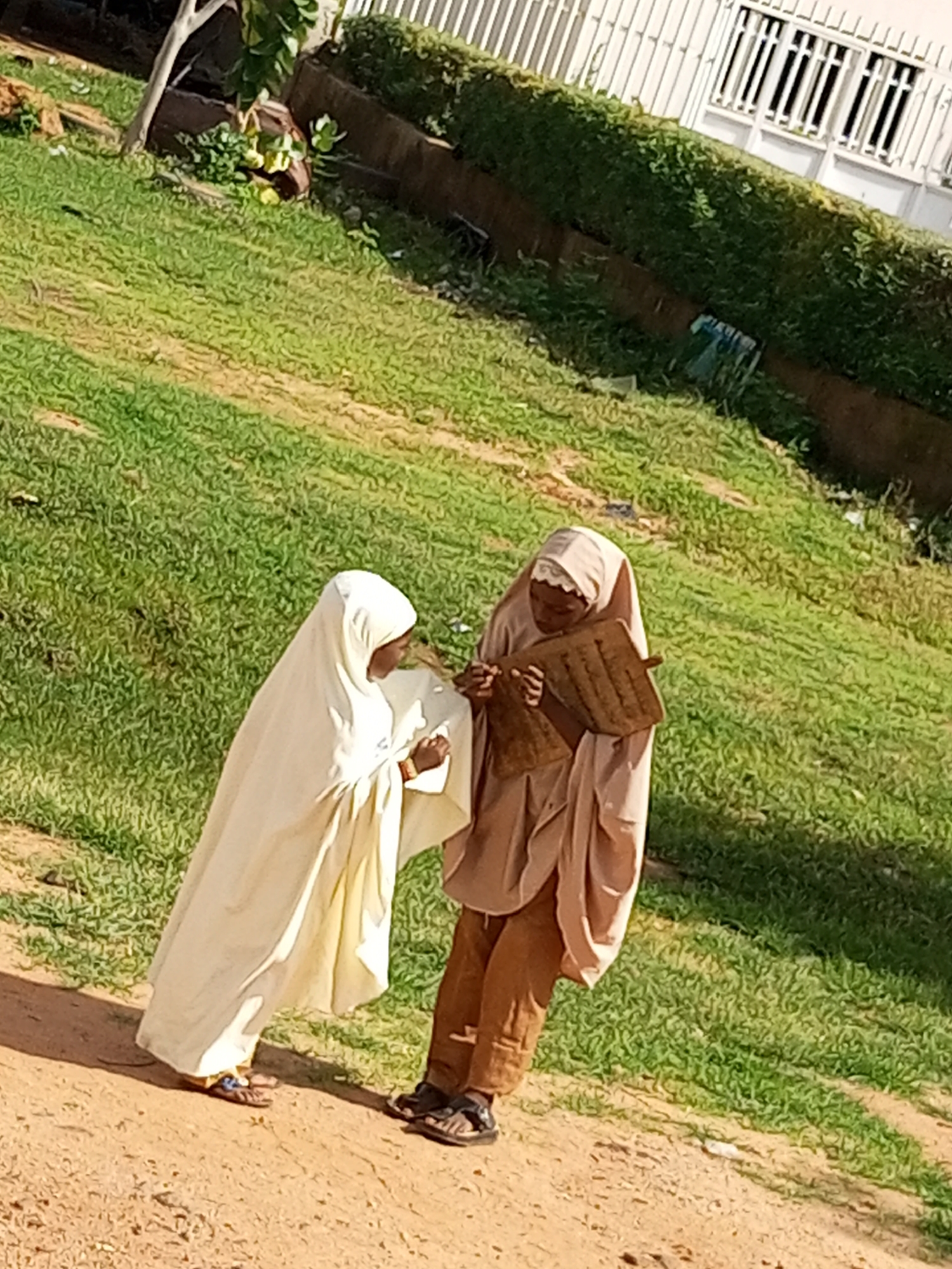 In Tumfure, Nigeria, two individuals are standing outdoors on a dirt path with green grass and a hedge in the background. One person, wearing a white hijab and a flowing white garment, is facing away from the camera and appears to be looking down. The other person, wearing a light brown hijab and brown pants, is facing the first person and holding a wooden tablet with Arabic script. The tablet is tilted towards the first person, suggesting they are interacting regarding its content. The environment is sunlit, with shadows cast on the ground, indicating daytime. A white fence and a building are visible behind the hedge.