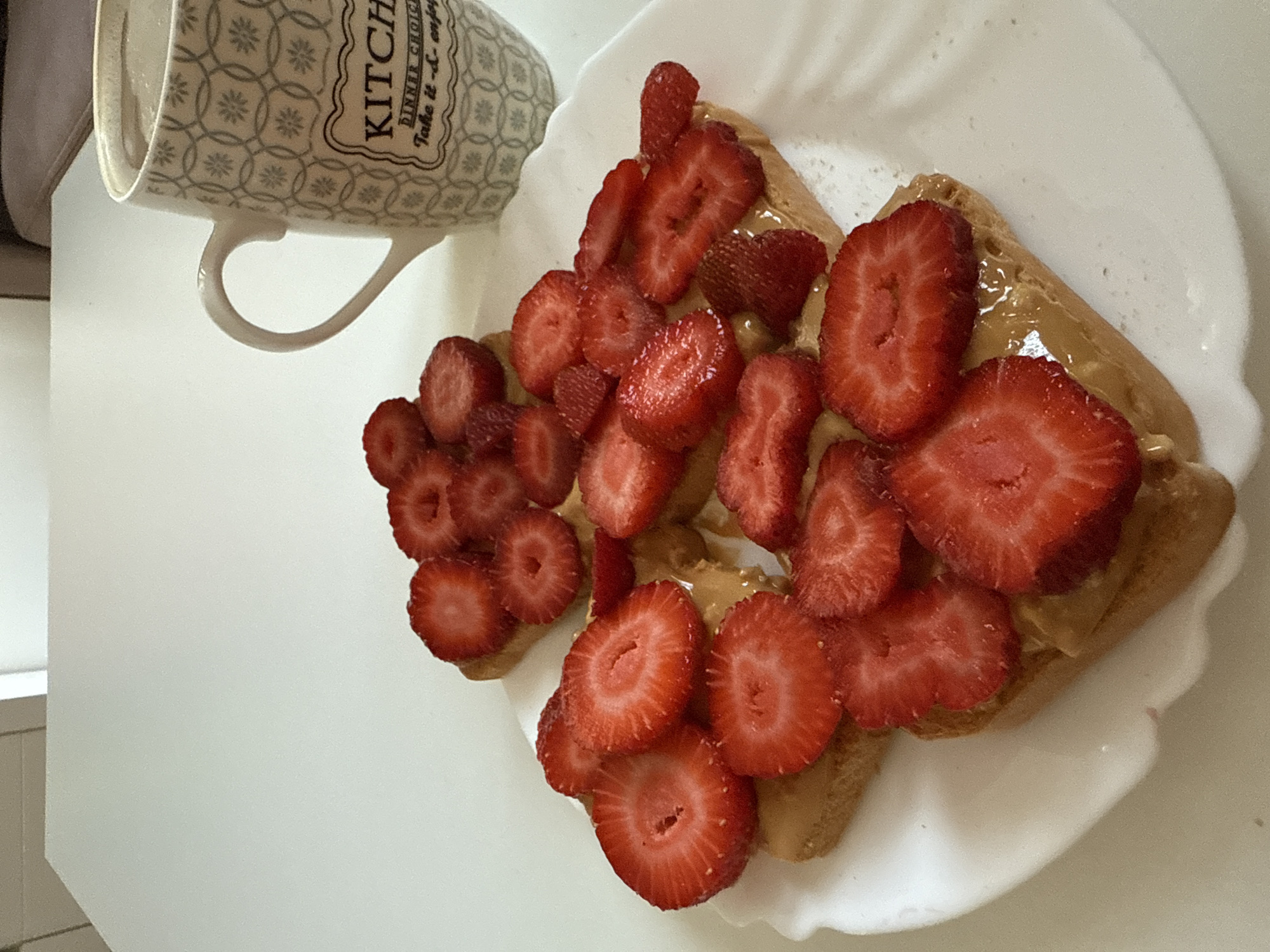 An overhead shot captures a white plate on a white table. On the plate are four pieces of toast, each topped with peanut butter and sliced strawberries. To the right of the plate, a white mug with a grey patterned design and the word "KITCHEN" printed on it is filled with a light-colored liquid. The background is blurred, showing a white surface and a hint of a chair. The scene appears to be indoors, possibly in a kitchen or dining area.