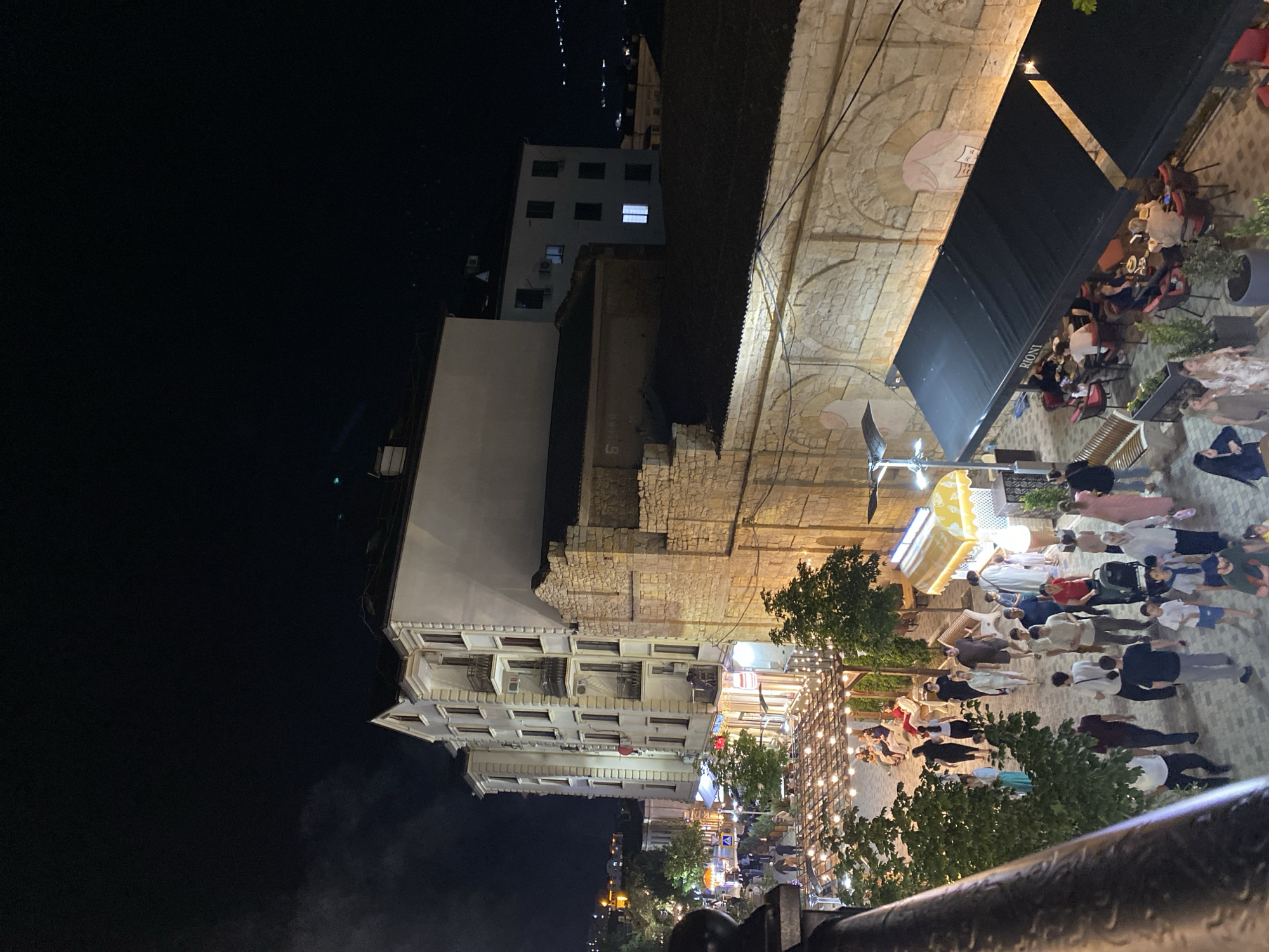 This is an eye-level, nighttime, aerial view of a city street in Derbent, Russia, likely taken from a balcony or upper floor. The scene is bustling with activity. A multi-story, cream-colored building with numerous balconies and windows dominates the left side of the frame, illuminated by interior lights. Adjacent to it is an older, rough-hewn stone building with arched openings.
The street below is paved with interlocking stones and is lined with trees and various establishments. A row of tables and chairs under black awnings, one bearing the name "RIONI," suggests an outdoor dining area for a restaurant or cafe. Many people are visible walking along the street and seated at the tables.
Streetlights and decorative string lights illuminate the area, creating a warm glow. Some people are dressed casually, while others appear to be dressed up for an evening out. The overall atmosphere is lively and social. In the background, more buildings and distant lights indicate the presence of a larger urban area. The night sky is dark, with no visible stars.