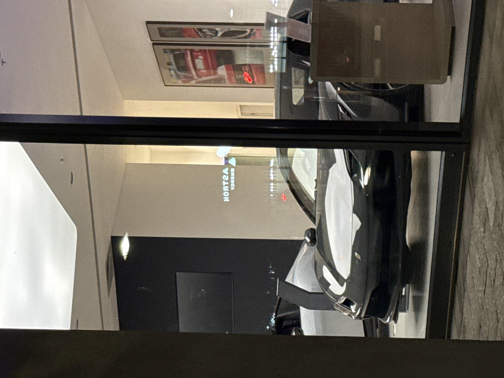 A black and white sports car is displayed behind a glass window. The car is parked on a showroom floor with a black wall and what appears to be a television. The ceiling of the room is white, and reflections on the glass show framed artwork. The reverse image of “ASTON ENERGY” is visible. The picture appears to be taken in a retail space in Cape Town, South Africa.