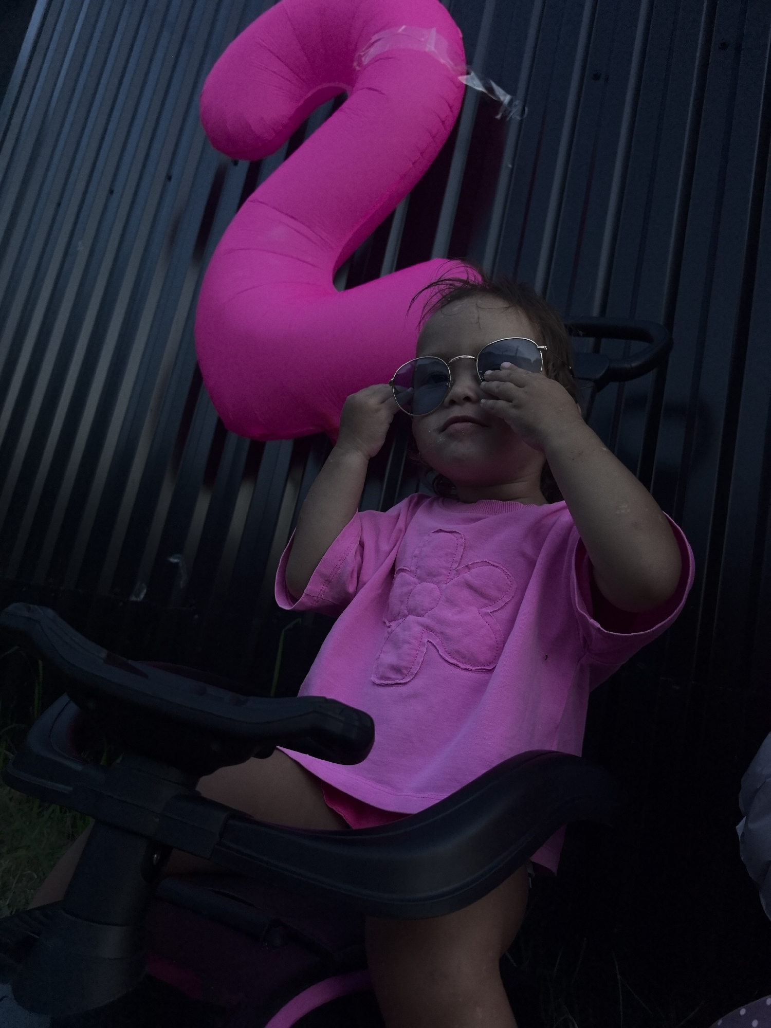 A young child wearing a pink t-shirt and sunglasses sits in a tricycle outdoors. Behind the child, a large, bright pink inflatable number "2" is visible against a dark, corrugated metal fence. The scene appears to be in an urban setting, possibly a yard or park in Chaikovsky, Russia. The lighting suggests it is either dusk or a dimly lit area.