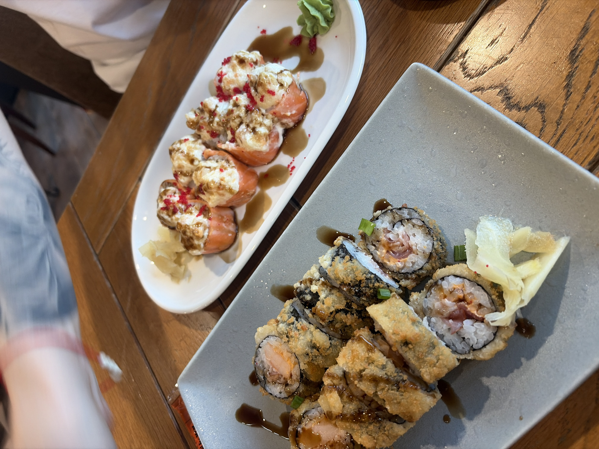 The scene depicts two plates of sushi rolls served on a warm-toned wooden table within a restaurant in Saint Petersburg, Russia. In the foreground, a rectangular grey plate holds several pieces of deep-fried or tempura sushi rolls, cut into segments, displaying a golden-brown crispy exterior, white rice, and a pinkish filling. These rolls are drizzled with a dark sauce, and some are garnished with chopped green onions. A portion of light-yellow pickled ginger is also present on this plate. In the background, an oval white plate contains eight pieces of baked or torched salmon rolls. Each roll is wrapped in salmon and topped with a creamy, browned sauce, garnished with small red sprinkles (likely tobiko). A dark sauce is drizzled over these rolls and on the plate, with a small dollop of green wasabi paste situated to the right. The top-left corner of the image shows a blurred arm and torso of a person wearing a light-colored top over a pinkish garment, suggesting movement. A second person's arm, clad in a white shirt, is partially visible in the top-right, indicating at least two diners. The wooden table exhibits a distinct grain pattern, and the background is softly blurred, revealing an indoor restaurant ambiance.