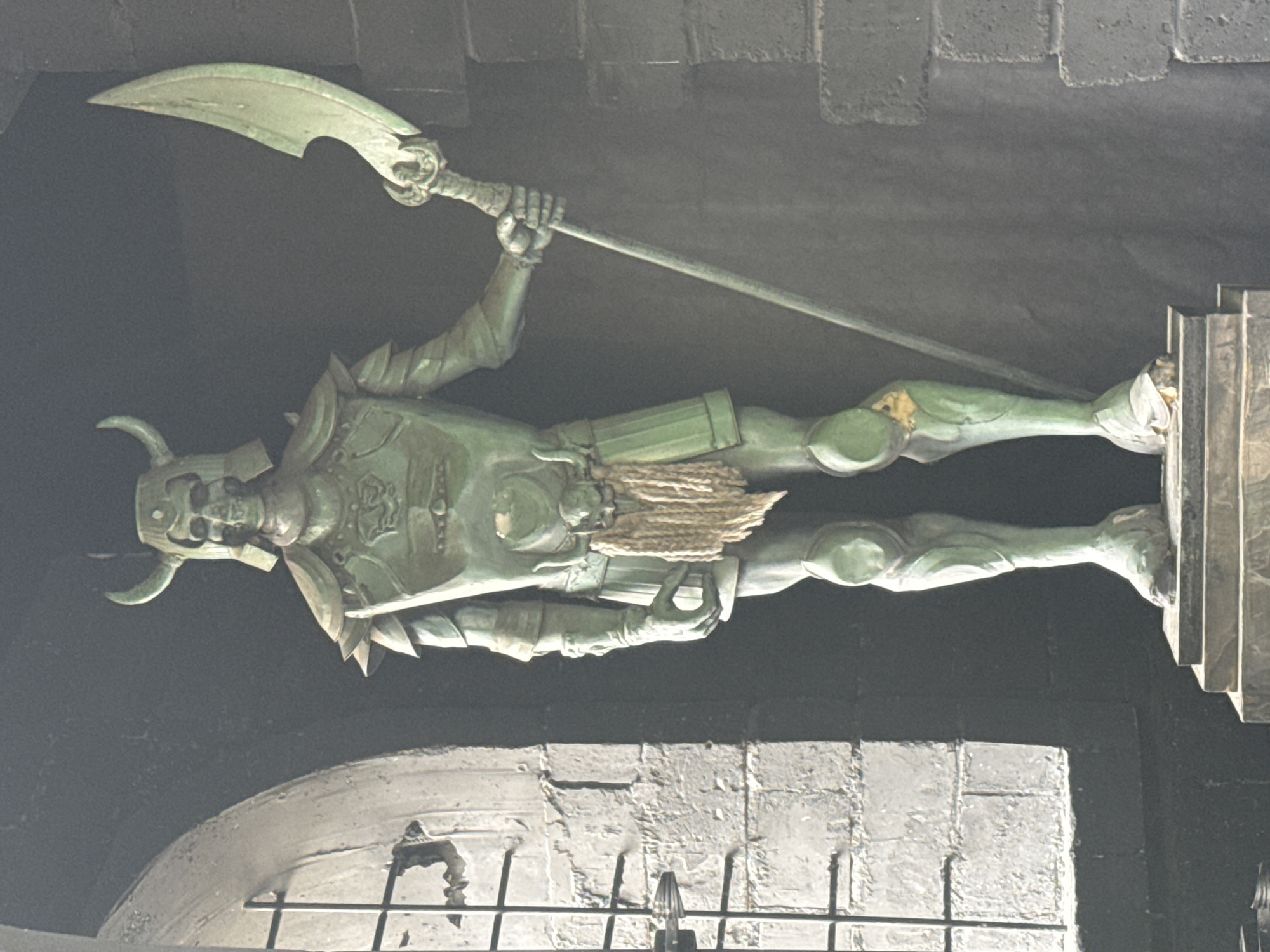 The image shows a bronze statue of a warrior figure with a horned helmet, holding a long polearm weapon. The statue is mounted on a rectangular plinth and is positioned above an arched entrance. The background appears to be a dark, possibly stone, structure. The photo was taken in the city of Yekaterinburg, Russia.