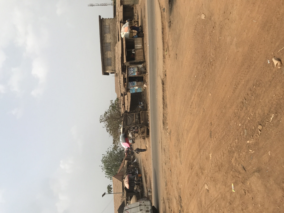 The image captures a bustling street scene in Ogbese, Nigeria, characterized by an unpaved reddish-brown earth foreground marked with tire tracks and minor debris. An asphalt road traverses the midground. Along both sides of this road, a series of single-story market stalls are visible, constructed with wooden frames and topped with corrugated metal roofs. Several stalls are shaded by open market umbrellas, featuring colors such as red and multi-striped patterns. On the right, at least two stalls bear prominent "POS" (Point of Sale) signage. Behind these stalls, a two-story concrete building with multiple windows is present, and further right, a metallic cellular communication tower is visible against the sky. On the left side of the road, a white commercial van is partially visible. Further back on the left, a solar-powered street light pole stands. Green trees are scattered throughout the background. Approximately five individuals are observed; one person walks centrally on the road section to the left, while others are situated near or within the market stalls, some appearing to be seated. The sky is overcast with a light grey hue, indicating daytime.