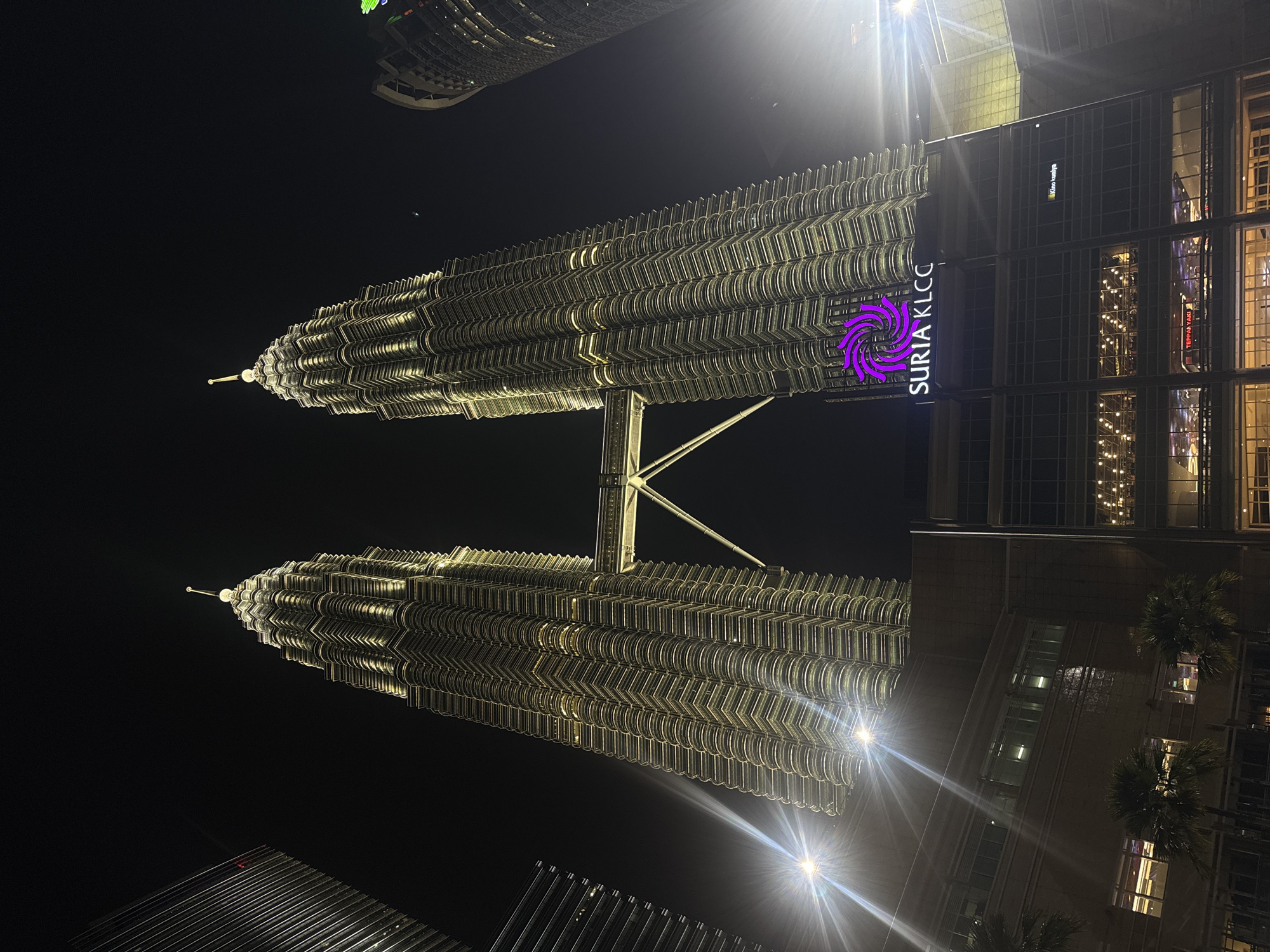 The photograph captures the Petronas Twin Towers in Kuala Lumpur, Malaysia at night. The towers are illuminated with a golden light, and the "Suria KLCC" sign is visible on a building adjacent to the towers. The sky is dark, and there are other buildings visible in the background. The towers are connected by a skybridge. Palm trees are visible in front of the adjacent building.