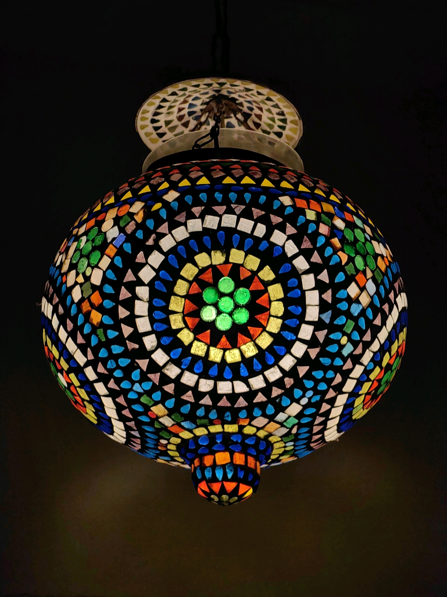 A Moroccan-style mosaic hanging lamp is the sole subject of this image, which is framed against a dark, indistinct background. The lamp has a spherical body adorned with a complex mosaic pattern of small, multicolored glass tiles. The tiles are arranged in concentric circular and radial patterns, with geometric shapes like squares and triangles. The dominant colors in the mosaic are blue, white, yellow, green, red, and orange, with a central circular section featuring bright green glass pieces. The lamp is suspended from above by a chain, and a soft light emanates from within, illuminating the mosaic from the inside and casting a warm glow on the dark background below. The lamp appears to be an interior decoration, possibly in a room or establishment. There is no discernible information about its location in Mumbai, India, as the image is devoid of any contextual clues like landmarks, street signs, or identifiable architectural features.