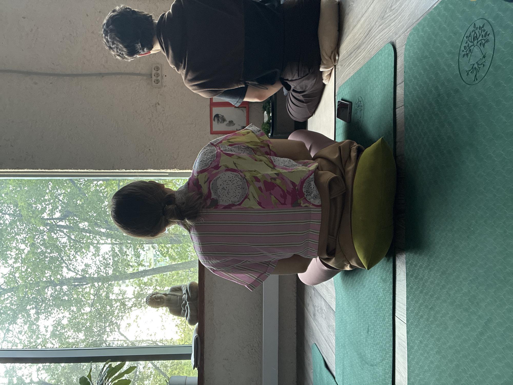 Two individuals are engaged in meditation in a room with an adjacent window offering a view of greenery, possibly trees, suggesting a location near a park or green space. The interior appears to be a studio or room dedicated to wellness practices.
The first individual, closer to the viewer, sits on a green yoga mat, using a green pillow for support. They are wearing pink leggings, a yellow shirt patterned with dragon fruit, and a pink and white striped shirt over it. Their hair is pulled back.
The second individual, positioned further back, is dressed in dark clothing. They also sit on a yoga mat, and a cell phone lies on the mat near them.
Visible near the window is a small statue of Buddha. The room has natural light, indicating it is daytime. The precise location within Moscow, Russia, cannot be determined solely from the visual information provided, but the environment suggests a quiet space suitable for meditation practices. Also visible are a framed picture on the wall and an electrical outlet above it.