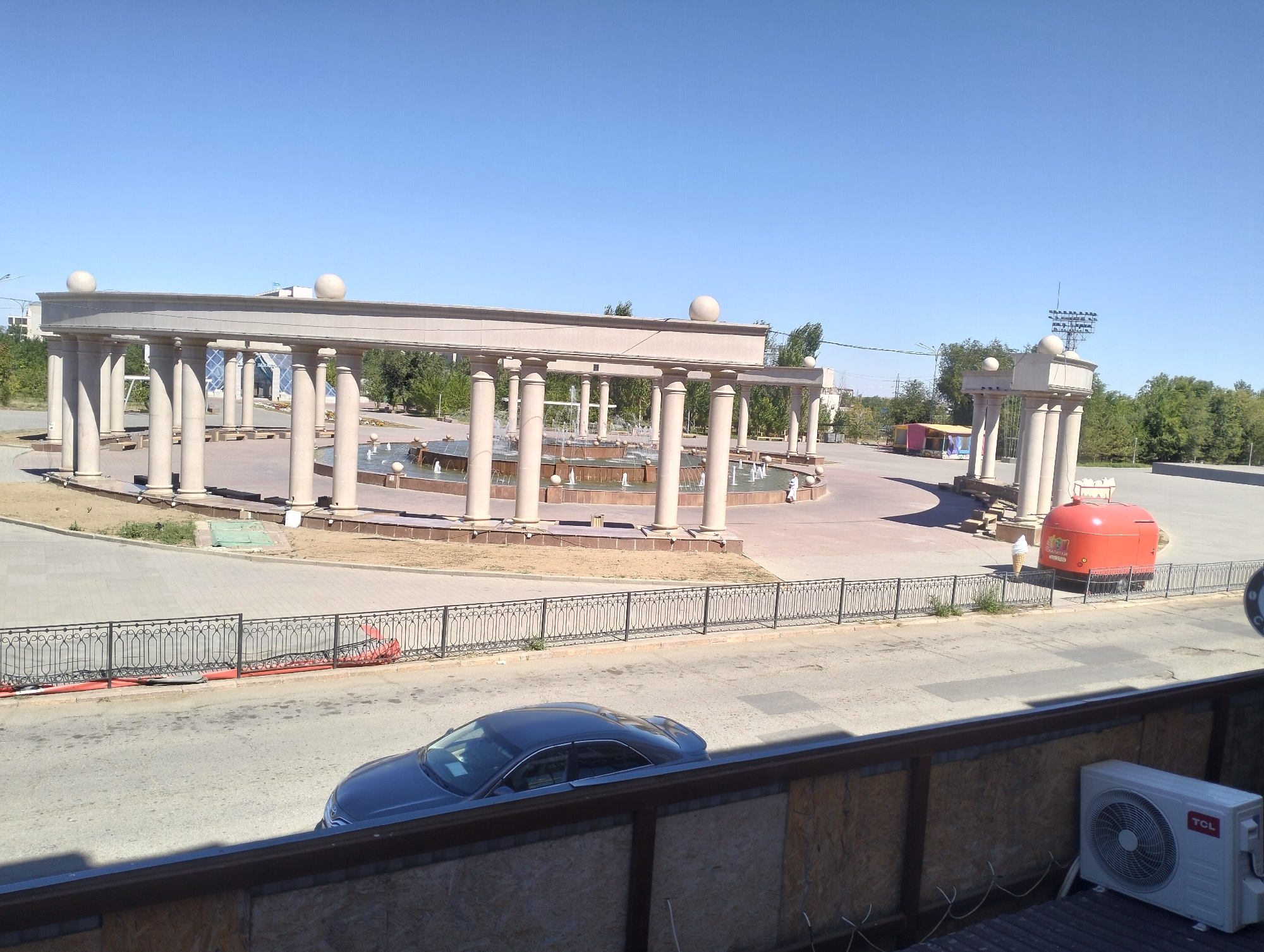 The image shows an outdoor scene in the city of Jezqazğan, Kazakhstan, on a clear day with a bright blue sky. In the foreground, a dark-colored sedan is parked on a paved road. A black metal fence runs parallel to the road. Behind the fence, a large, open-air structure featuring numerous tall, light-colored columns supporting an overhead walkway is visible. This structure appears to be part of a public space or monument, with fountains operating within its central area. Further in the background, green trees and a light-colored building with a modern architectural style can be seen. To the right of the columned structure, a bright red, cylindrical vehicle, possibly a food cart or small bus, is parked. An ice cream cone is placed on the ground near the red vehicle. An outdoor air conditioning unit is mounted on a wooden surface in the lower right corner of the image.