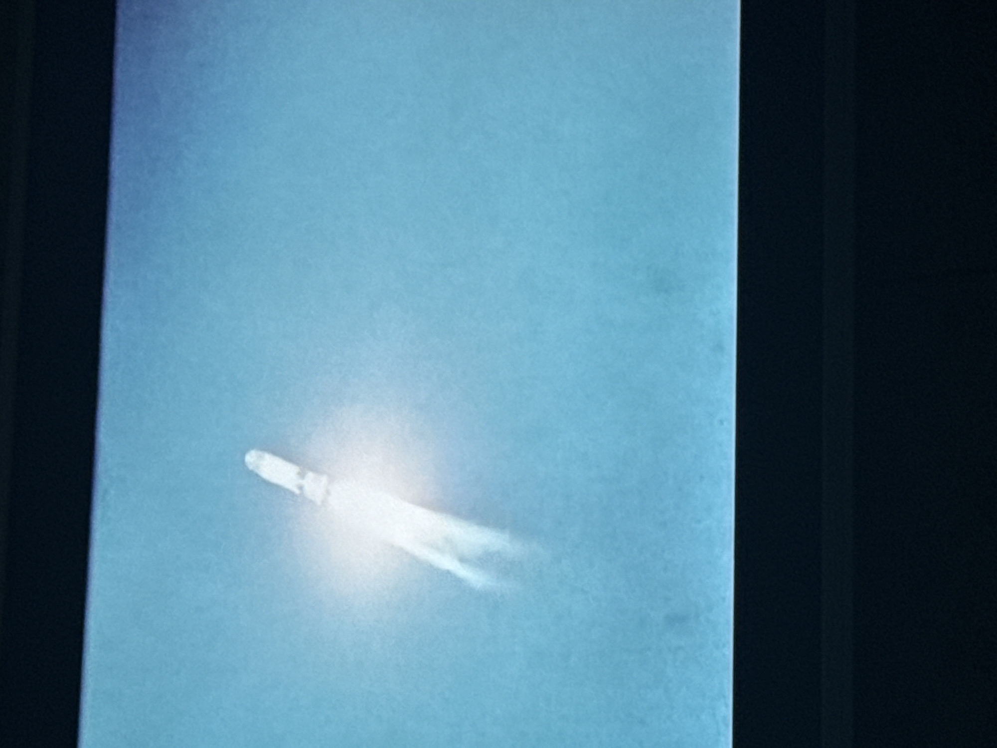A photograph shows a rocket in flight against a blue sky. The rocket is emitting a trail of exhaust. The photograph appears to be of a projection on a screen, as evidenced by the screen's frame visible on the sides of the image. There are no people visible in the photograph. The location of Mytishchi, Russia is not directly related to the content of this image.