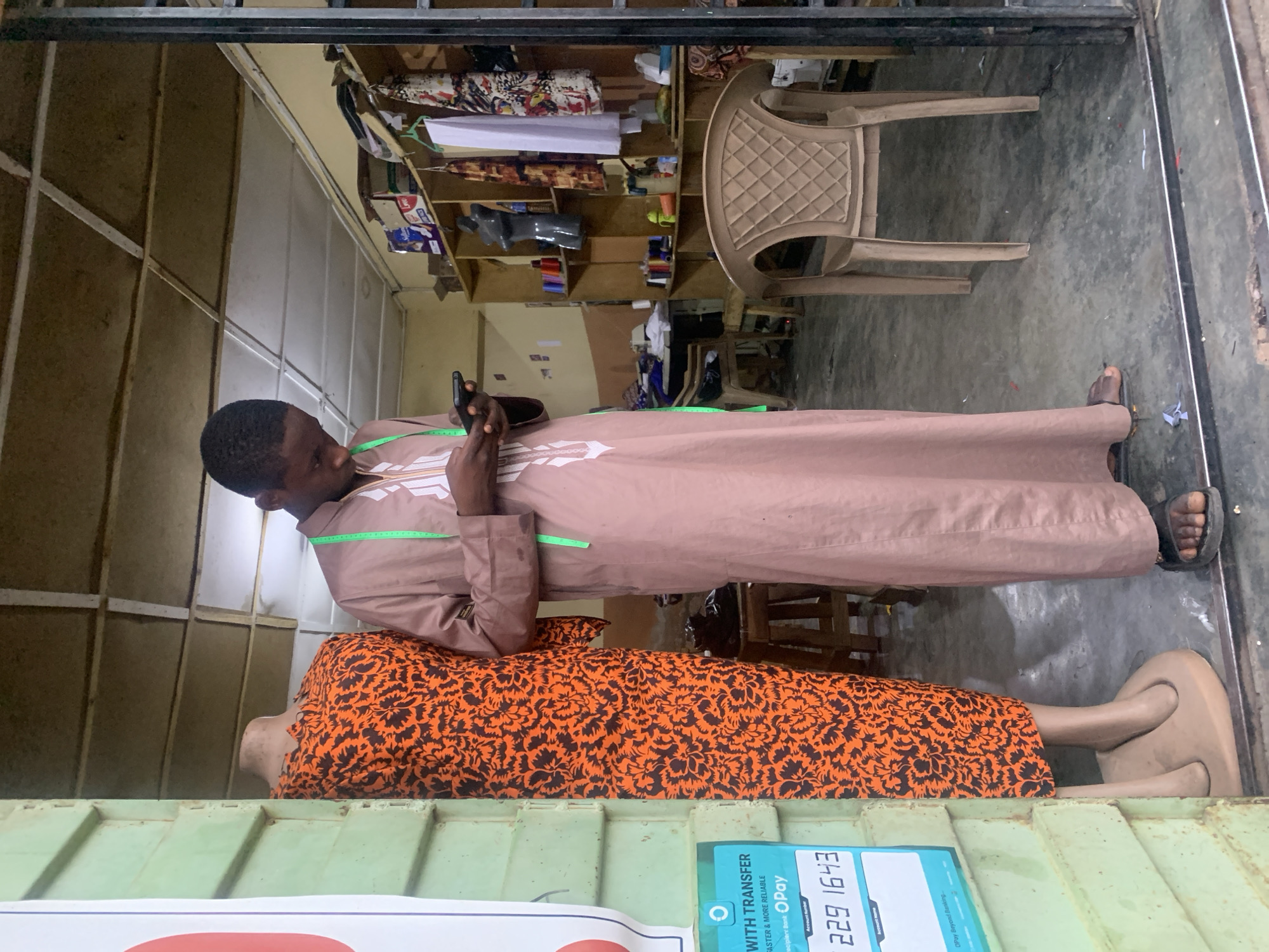 A young man, likely a tailor, stands in what appears to be a small tailoring or fabric shop, looking at his mobile phone. He is positioned next to a mannequin that is dressed in a vibrant orange dress with a black floral or leaf pattern. The man wears a long, light brown robe-like garment adorned with white embroidered patterns and has a green measuring tape draped around his neck, indicating his profession. He is wearing sandals.
The shop interior features simple wooden shelves against light-colored walls, stocked with various fabrics, spools of thread, and other supplies. A light-colored plastic chair is visible further back in the shop. The floor is a plain, possibly concrete, surface. The overall setting suggests a bustling, functional workspace, common for small businesses in a location like Jos, Nigeria.
In the foreground, partially visible at the bottom of the image, is a blue and white sign. Visible text on this sign includes "WITH TRANSFER", the logo for "OPay", and an "Account Number: 229 1643". Other smaller text like "FASTER & MORE RELIABLE" is also legible. The time of day appears to be daytime, with general ambient light filling the shop. No specific emotions are discernible from the man, who seems focused on his device.