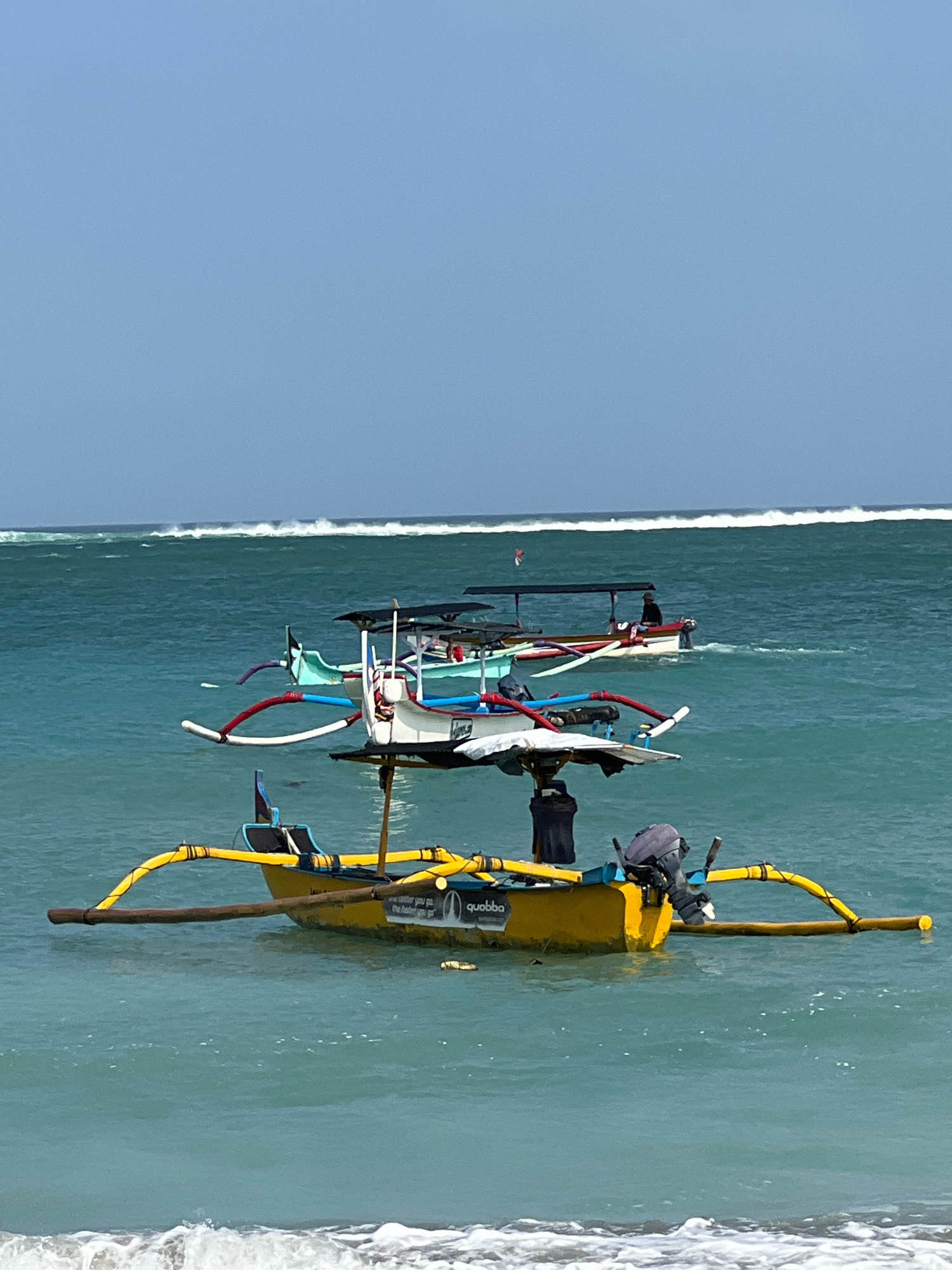 In Kuta, Indonesia, several traditional outrigger boats are anchored in the turquoise ocean under a clear blue sky. The water is calm with gentle waves rolling in from the horizon. The foreground features a bright yellow boat with prominent yellow outriggers and a small outboard motor. This boat has some text on its side, including the word "quobba." Behind it, other boats of various colors, including white, light blue, and red accents, are visible. One boat has a person sitting on it, appearing to operate the motor and a red flag is flying from a pole. In the distance, a long line of white-capped waves indicates a significant surf break further out at sea. The scene is one of serene ocean activity characteristic of a popular beach destination.