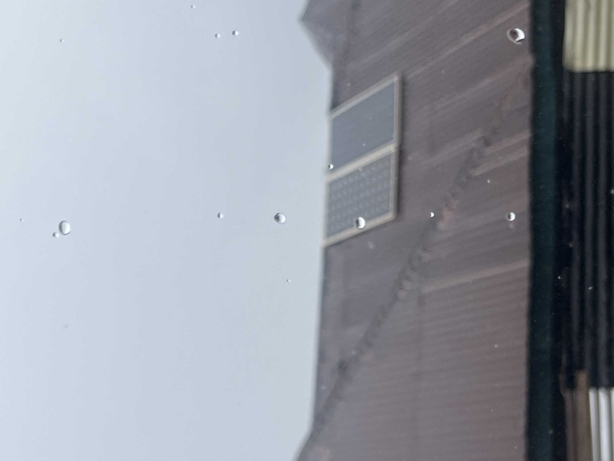 The image is a close-up, abstract shot featuring water droplets on a glass surface. The foreground is dominated by a light, overcast sky filled with numerous small, clear water droplets. Some droplets are perfectly round, while others are elongated or irregular shapes, appearing to be in motion or recently settled. A few larger, more prominent droplets are visible, reflecting light and distorting the background.
In the background, a portion of a building is visible, blurred and out of focus due to the shallow depth of field. The building appears to be made of dark, textured material, possibly wood or a dark-colored siding, with horizontal lines suggesting a ribbed texture. There are two square or rectangular panels on the building's facade, one above the other, which appear darker and smoother than the surrounding material. These panels might be windows or decorative elements. To the right of the building, a section of vertical blinds or a similar striped facade is visible, also out of focus.
The overall impression is one of a rainy or dewy day, with the camera positioned behind a wet window or surface looking out at a building. The lighting is diffused and muted, consistent with an overcast sky. There is no visible text in the image. The focus is on the textures and light play of the water droplets against the muted, out-of-focus background. The scene evokes a sense of stillness and quiet, perhaps a moment of contemplation on a damp day. The location context of Gembu, Nigeria, suggests a possibly tropical or subtropical climate where such weather conditions might occur.