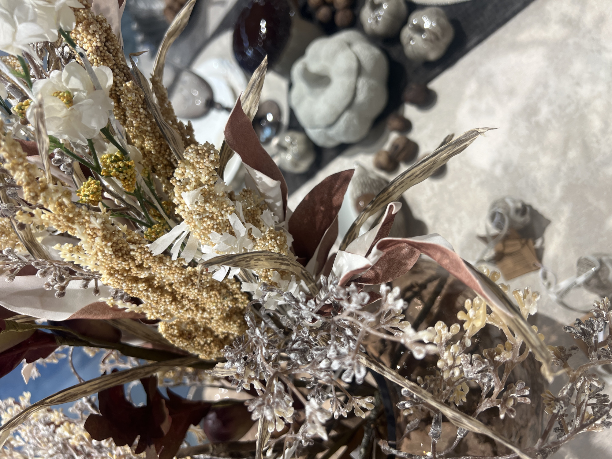 The image shows a close-up of a dried floral arrangement, likely a fall or autumn display. The arrangement features various textures and colors, including dried grasses, small beaded seed pods, and some feathery elements that appear to be dusted with white or silver. There are also some deep red leaves or flower petals interspersed. In the background, out of focus, are several small decorative pumpkins or gourds, some of which are white or light gray, and a dark, glossy object that might be another decorative item or a piece of fruit. The overall impression is of a still life, possibly a table setting or windowsill decoration. The lighting suggests an indoor environment, possibly with natural light coming from a window. The scene does not contain any identifiable landmarks or elements that would place it specifically in Moscow, Russia.