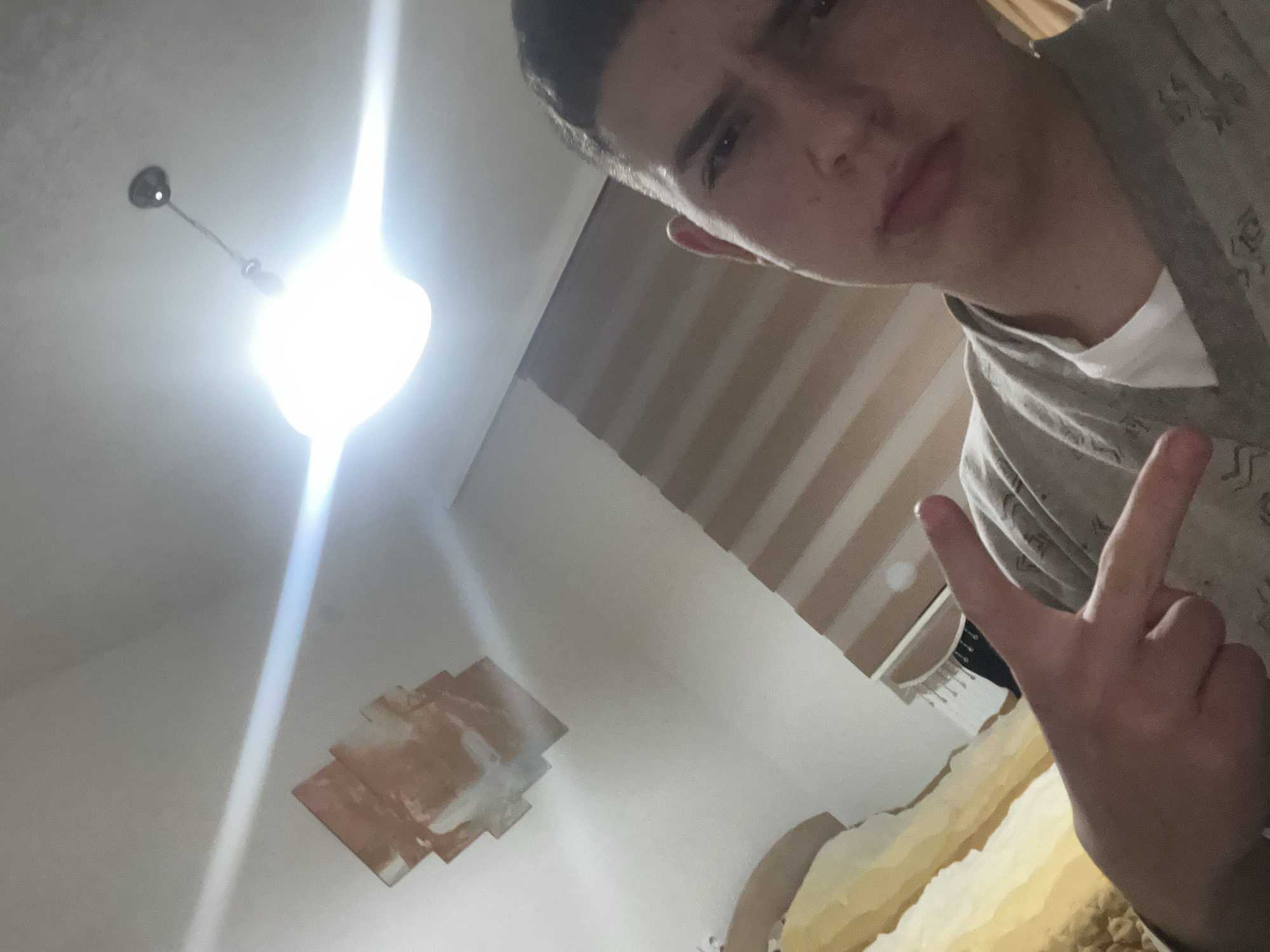 A selfie-style, indoor photograph shows a young Caucasian male, presumably in Zlatograd, Bulgaria, though there is no confirming evidence. The male presents from the upper right of the frame and is angled downward. He is wearing a light-colored shirt under a button-up garment. His right hand is raised, displaying a "V" gesture. The background includes a ceiling with a light fixture emitting intense light. A wall with some architectural detail or repair work is visible, as is a bed. Striped wallpaper is visible in the upper right portion of the image. The overall lighting conditions appear to be bright.