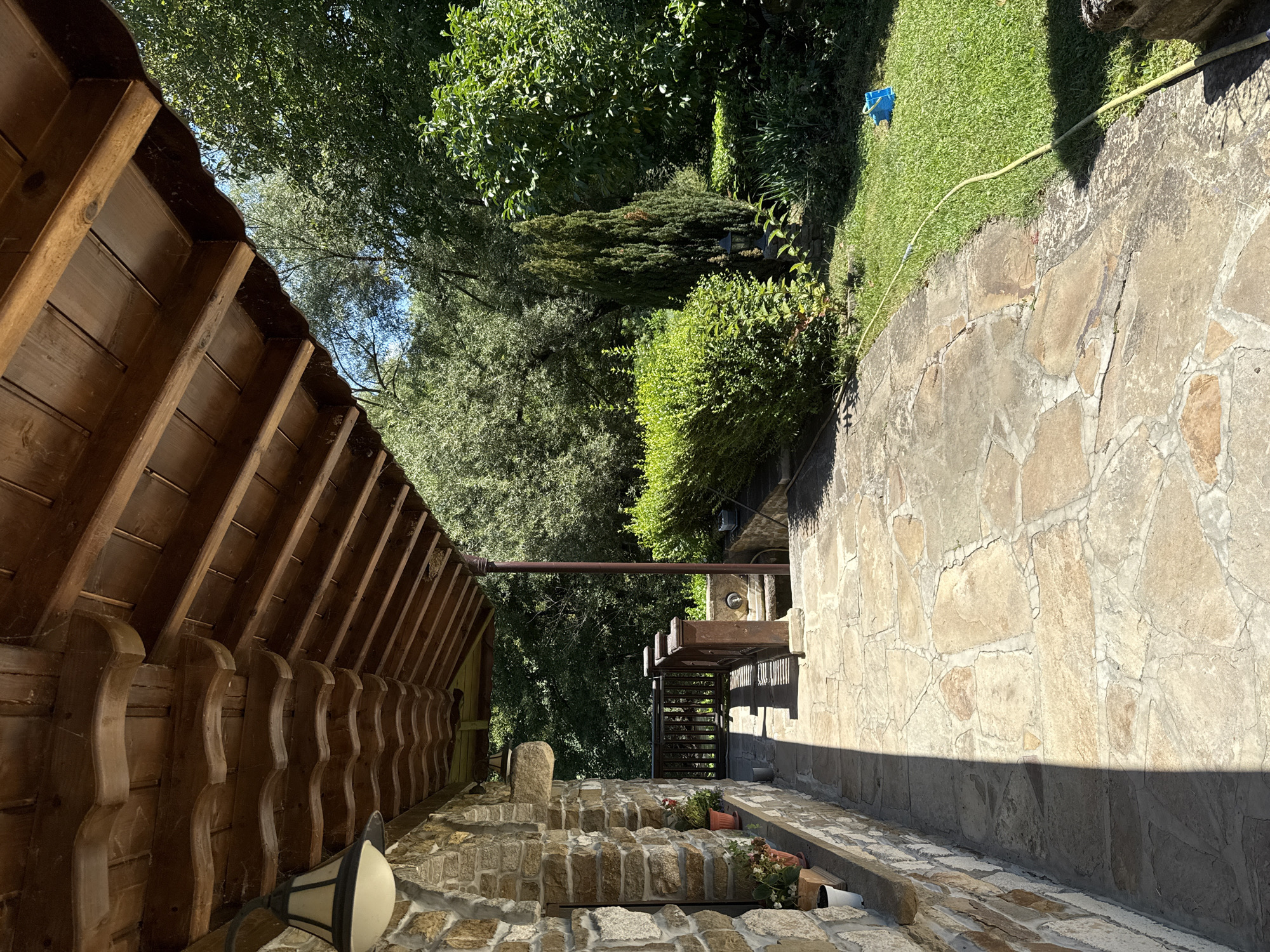 The image depicts a sunlit stone patio leading away from a rustic stone building with a wooden overhang. The overhang has ornate wooden detailing and supports. A stone wall of the building is visible on the left, with several potted plants placed on a ledge. A wooden railing with posts is situated further down the patio, separating it from a lower area. Beyond the railing, lush green trees and bushes create a natural backdrop. A narrow, conical evergreen tree stands prominently among the foliage, and a garden hose is visible on the grass to the right, with a blue object near its end. The scene appears to be a private outdoor space, possibly a garden or yard, with a distinctly rural or traditional architectural style. The location is Petrevene, Bulgaria.
