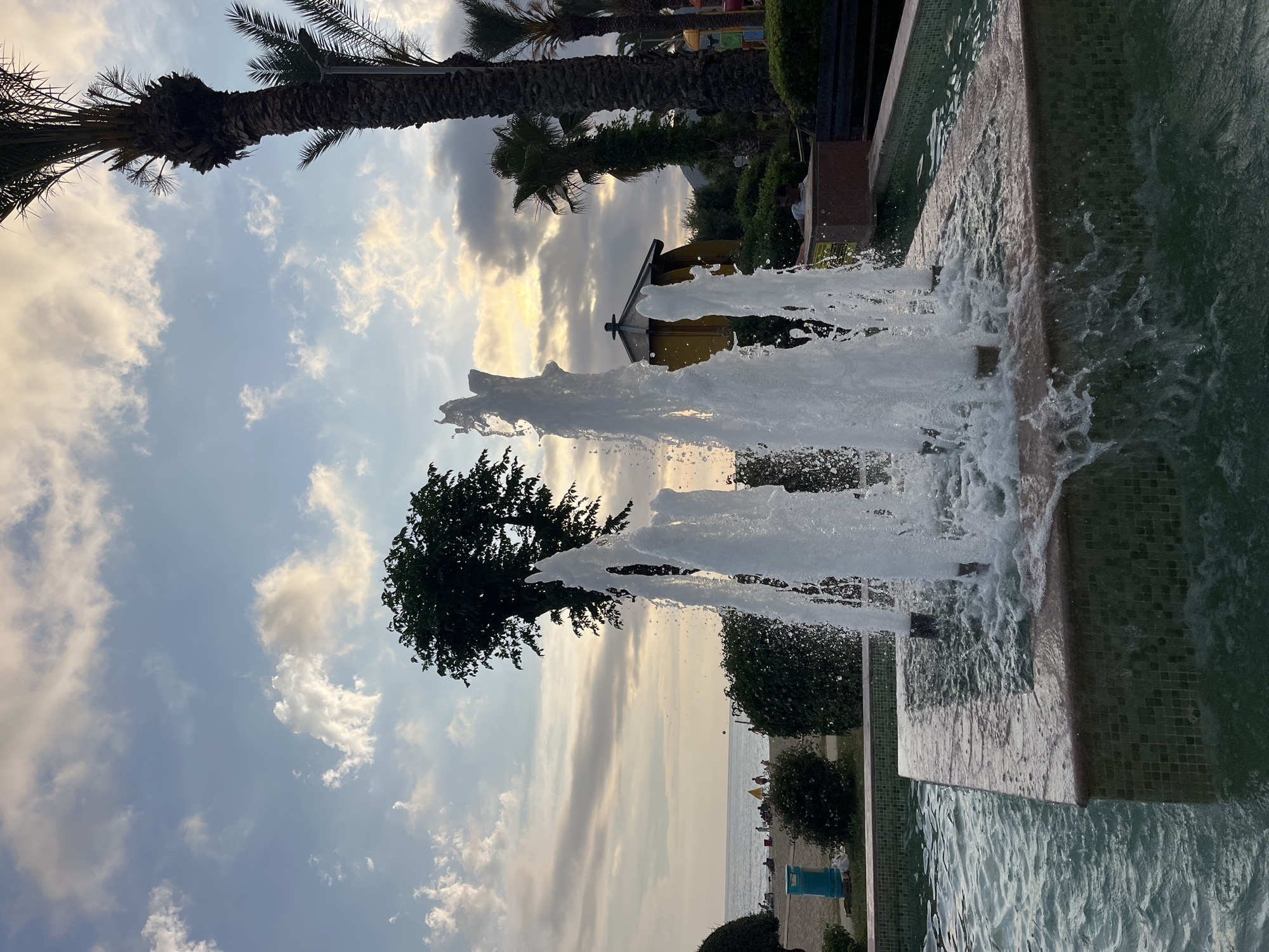 The image shows a water fountain at a beach in Alanya, Turkey. The sky is partially cloudy with the sun setting in the background. There are palm trees in the background, along with a few structures. The water fountain is the main focus, with water jets spraying upwards. The fountain is built with tiles. In the background, beachgoers can be seen near the sea.
