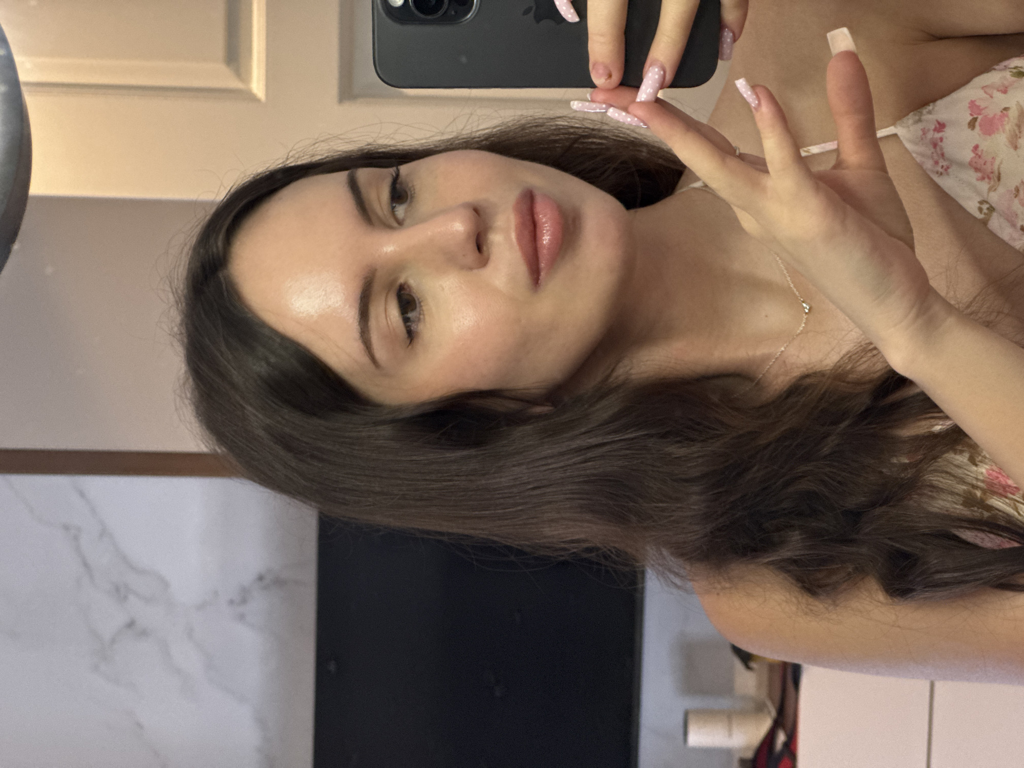 A woman with long, dark brown hair is taking a selfie in a mirror. She is looking at the camera and her face is partially obscured by her hand, which is holding her phone. She is wearing a light-colored, floral-print top. Her fingernails are long and painted pink with white polka dots. A delicate necklace with a small pendant is visible around her neck. The background appears to be an indoor setting with a marble-patterned wall and what looks like a door in the background.