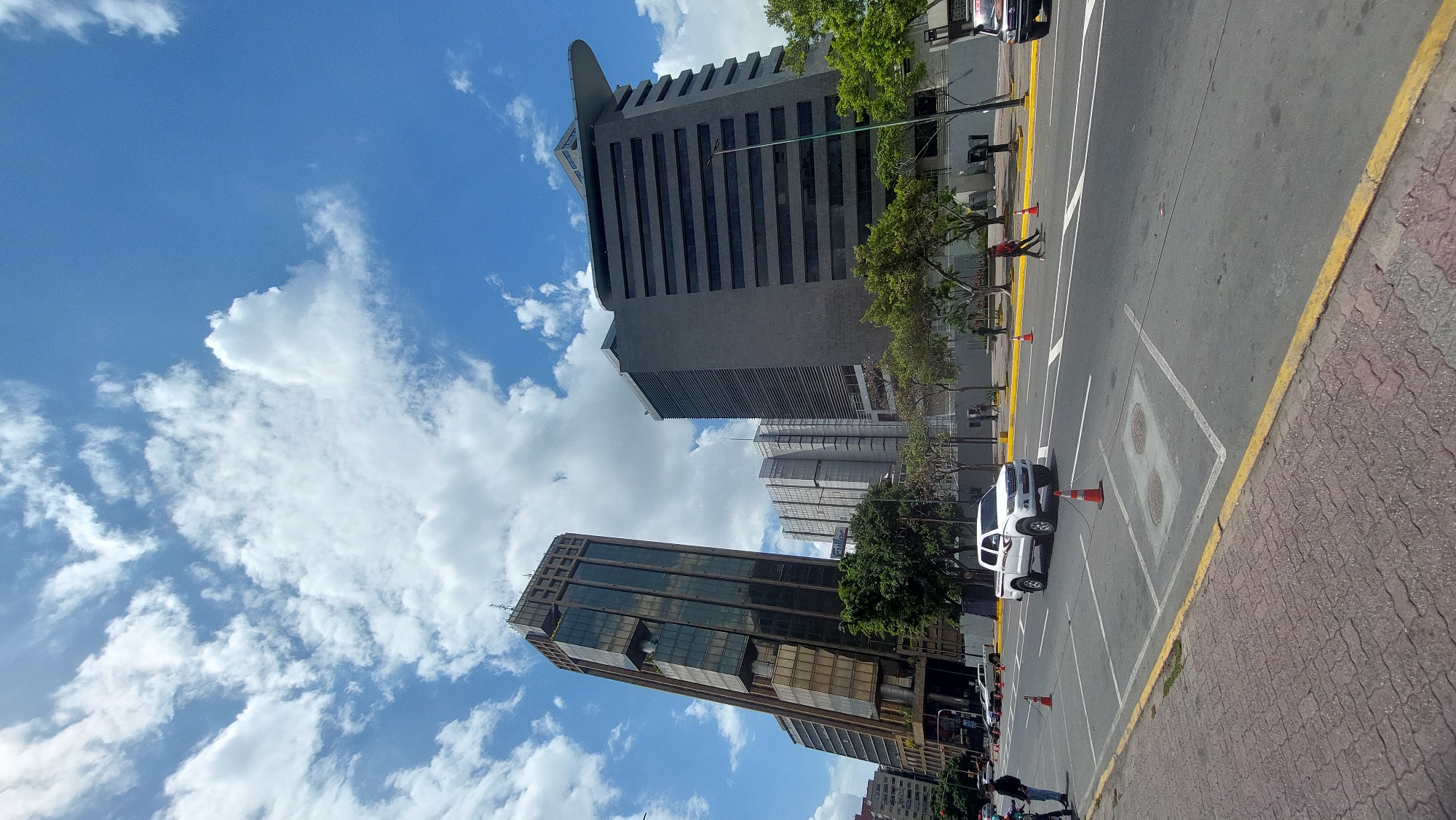 The image captures an urban scene in Caracas, Venezuela. The perspective is angled upward, showcasing a street lined with modern buildings under a sky filled with scattered clouds. Two prominent skyscrapers dominate the view, one with a reflective glass facade and the other with a gray, geometric design. A street runs alongside the buildings, marked with yellow and white lines and several orange traffic cones. A white vehicle is parked within the designated lines, and several other cars are parked further down the street. Pedestrians are visible on the sidewalk. The overall environment suggests a busy commercial district within the city.