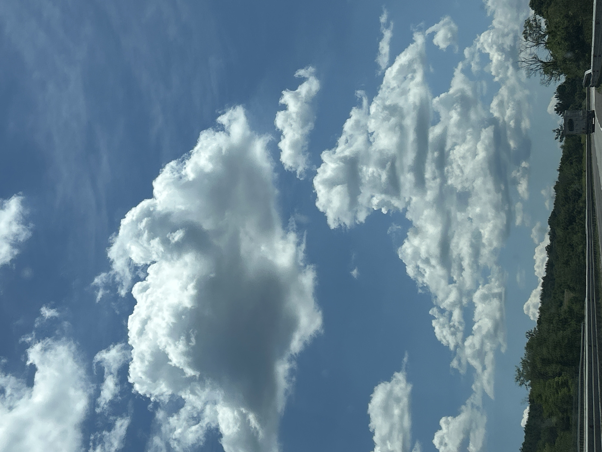 The image captures a bright, partly cloudy day over a highway, possibly near Verinsko, Bulgaria. The sky is a vibrant blue, dotted with large, fluffy white cumulus clouds. Below the sky, a dense, tree-covered hillside forms a natural backdrop. A highway runs horizontally across the lower portion of the image, with a concrete barrier and guardrail visible along its edge. A single vehicle, a light-colored truck, is seen driving on the highway. The overall scene suggests a rural or semi-rural setting, with a focus on the natural landscape and the open road.