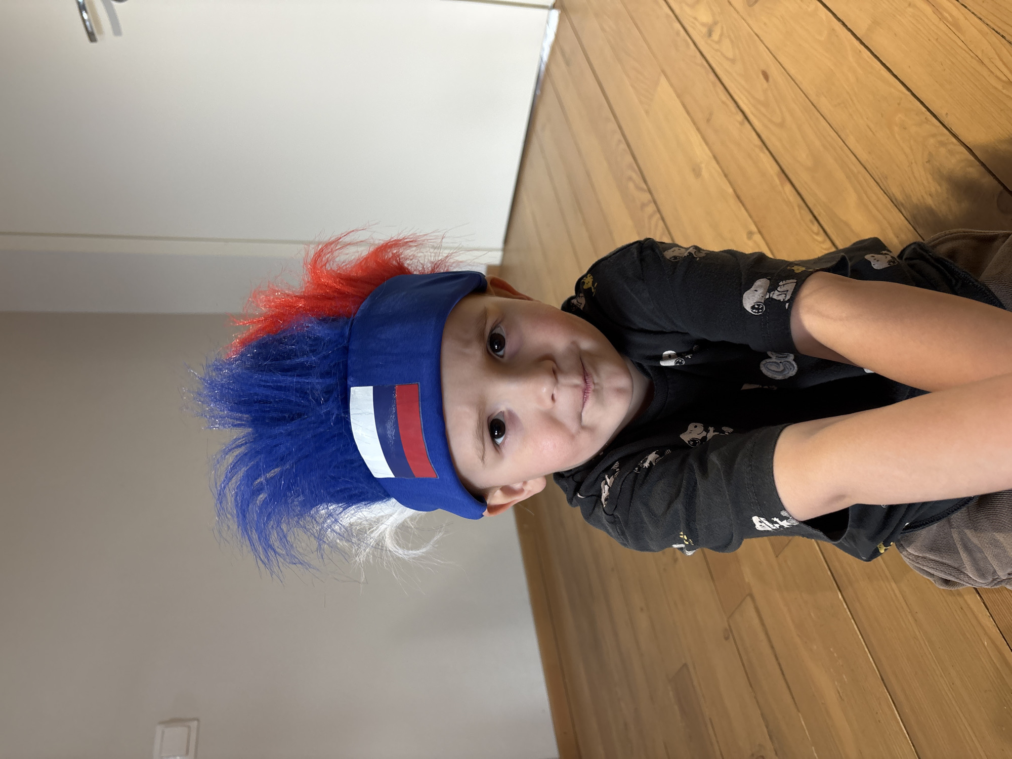 A young child wearing a novelty hat with red, white, and blue faux hair lies on a wooden floor. The hat is a blue headband with vertical stripes of white and red on one side. The child is wearing a black t-shirt with Snoopy cartoon characters. The setting appears to be indoors, with a white door visible in the background. There is no explicit information present in the image to directly confirm the location in the city of Chaikovsky, Russia.