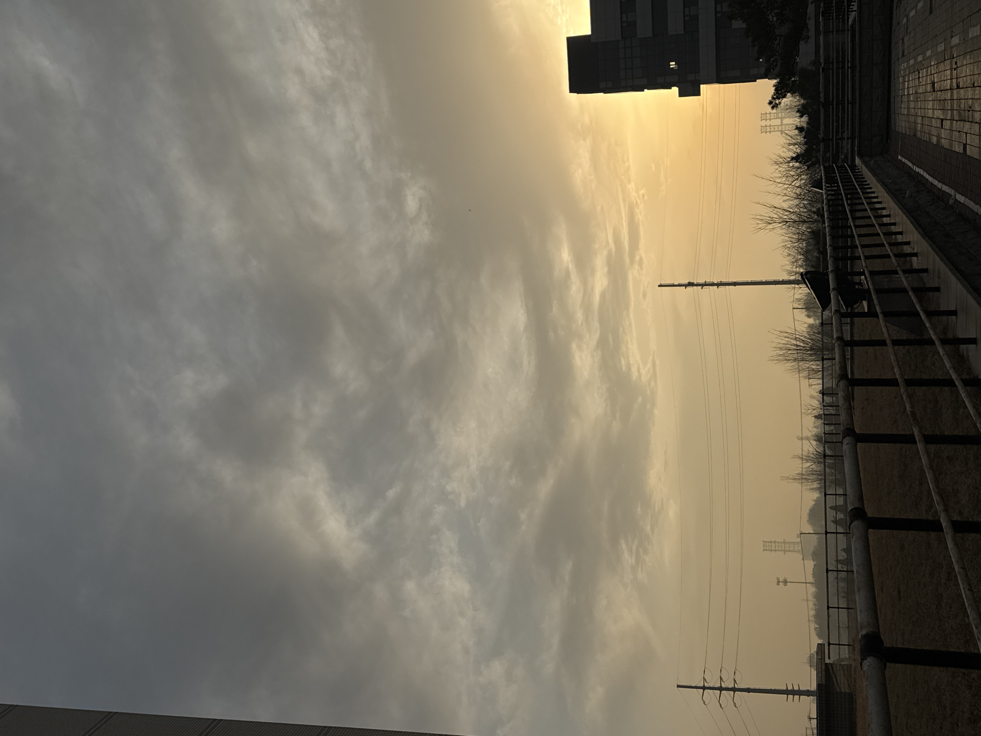 The image shows an overcast sky with a yellowish-orange tint, potentially indicating sunset or air pollution. The scene is located in an urban area, likely Zhongtun, China, based on the prompt. On the right side of the image, a walkway with a railing runs along a raised area. Power lines stretch across the yellowish-orange sky. A building is visible at the top of the image.