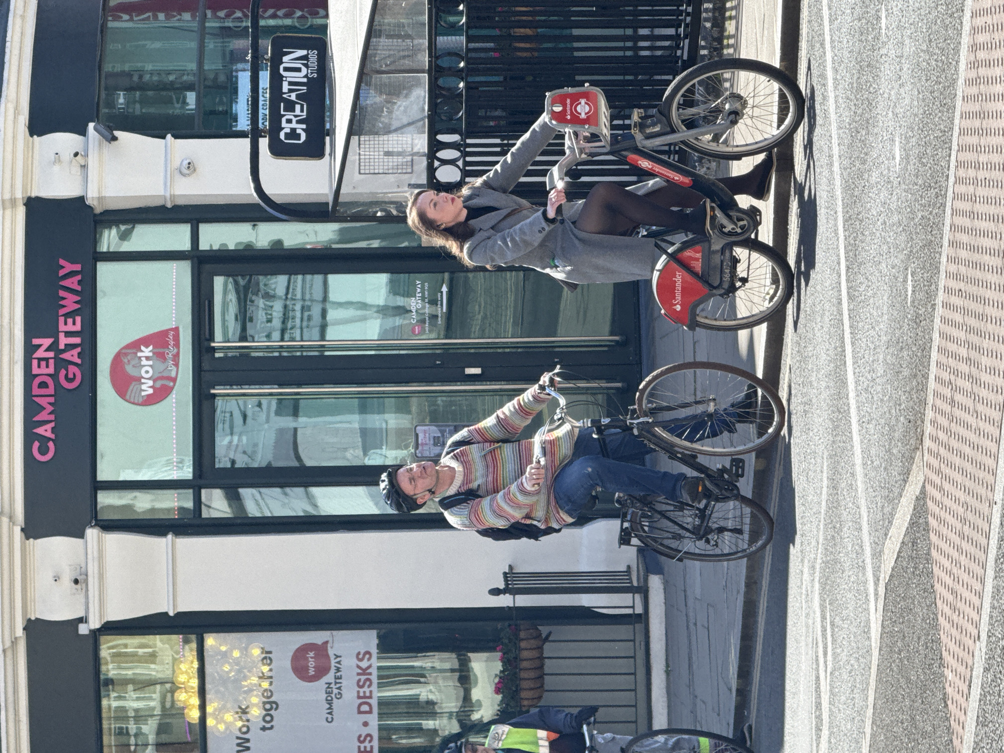 The image shows two people riding bicycles on a city street in London, United Kingdom. A woman is on a red Santander bike, wearing a gray coat, black top, and tights. A man is on a black bike and is wearing a helmet, a striped sweater, a backpack, and jeans. In the background, there is a building with the words "CAMDEN GATEWAY" and "CREATION STUDIOS" visible on signs. The street is paved, and there is a sidewalk on the right side of the image. A road with white traffic lane dividers is visible behind the pedestrians.