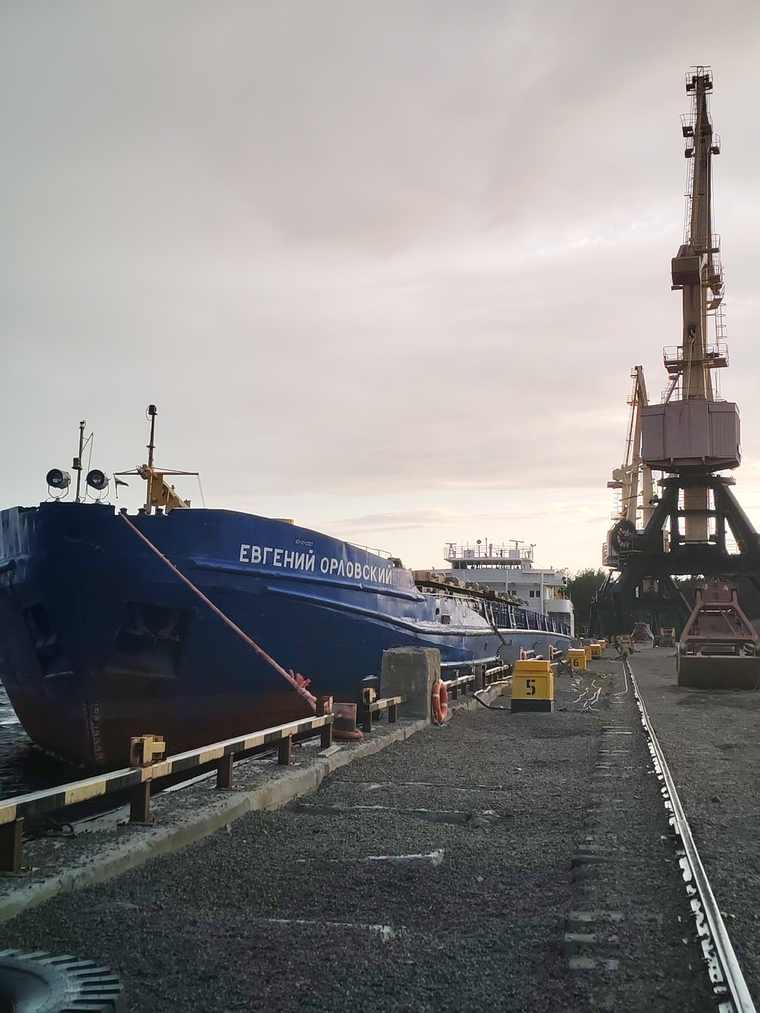 The image depicts a port scene in Shal'skii, Russia, under an overcast sky. In the foreground, a large blue cargo ship named "ЕВГЕНИЙ ОРЛОВСКИЙ" is docked alongside a pier. The ship is secured by a thick red rope. Adjacent to the ship are railway tracks laid on the ground, which is covered in gravel. Several yellow rectangular markers with numbers "5" and "6" are placed along the tracks. Two large cranes, one more prominent than the other, are visible in the background, suggesting loading or unloading operations. The environment appears industrial, with the water of the port visible on the left. There are no people visible in the frame. The lighting suggests it might be early morning or late afternoon.