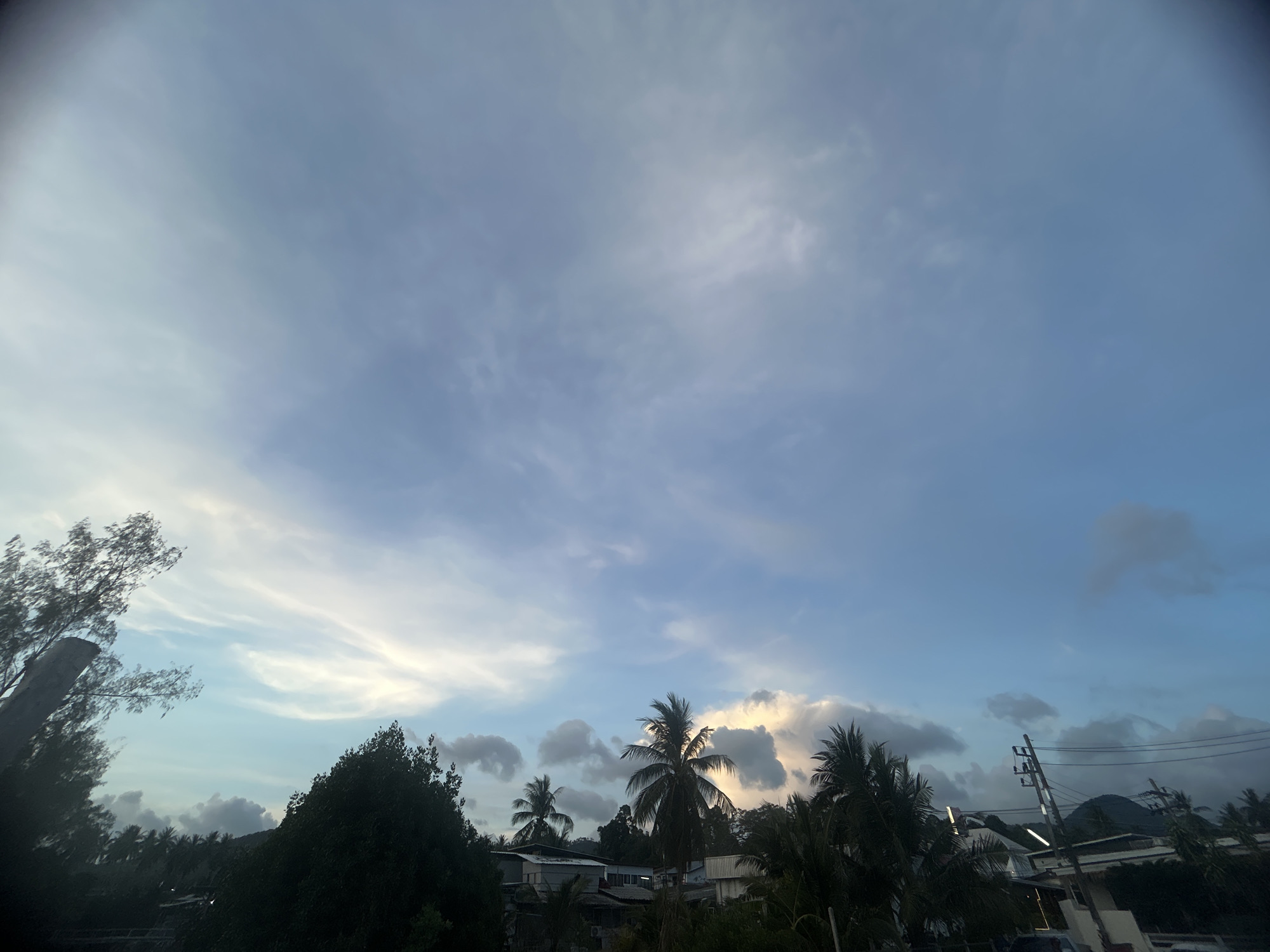 The image shows a daytime scene in Baan Sri Thanu, Thailand. The sky is mostly blue with scattered white and grey clouds. Palm trees and other vegetation are visible in the foreground, partially obscuring buildings and infrastructure. The scene suggests a tropical or coastal environment.