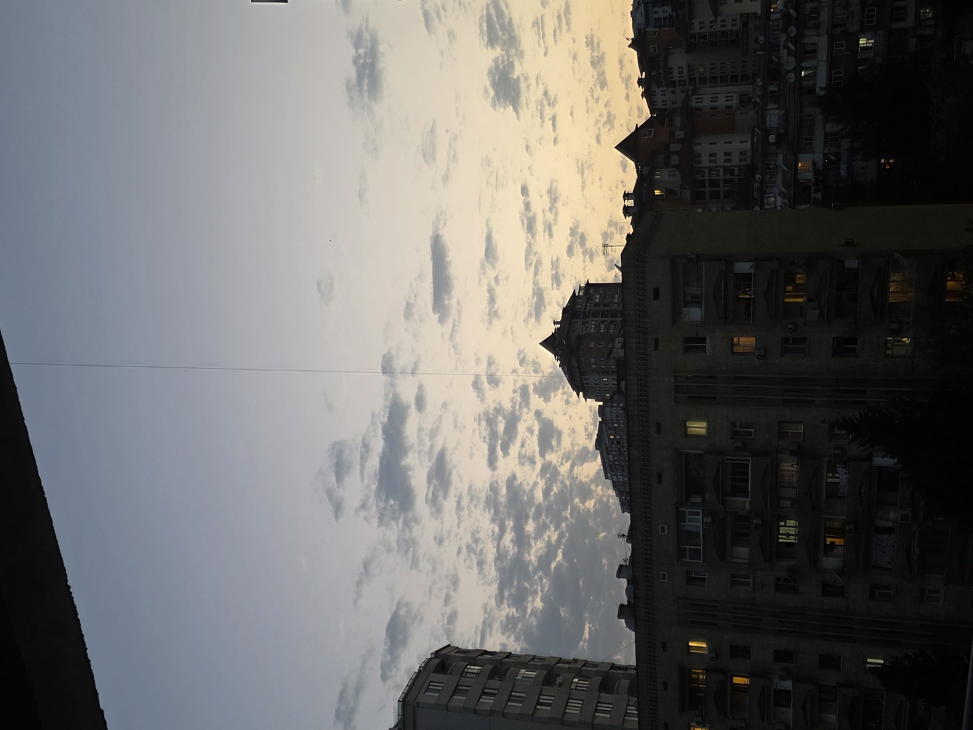 The image depicts an urban landscape at dusk, likely in Baku, Azerbaijan, as suggested by the architectural style of the buildings. A string is visible dividing the sky vertically, possibly a wire or cable. The sky is a gradient of twilight colors, transitioning from a pale blue at the top to a soft yellow and orange near the horizon, with scattered clouds. In the foreground, the silhouettes of several apartment buildings are prominent. The closest building on the left is a tall, modern-style apartment complex with a curved facade and numerous windows, some of which are illuminated, indicating occupancy. To its right and in the background, older, more densely packed residential buildings with varied rooflines, chimneys, and architectural details are visible. Some of these buildings also have illuminated windows. The overall scene conveys a sense of evening in a residential urban area, characterized by the interplay of natural light and artificial illumination from the apartments.