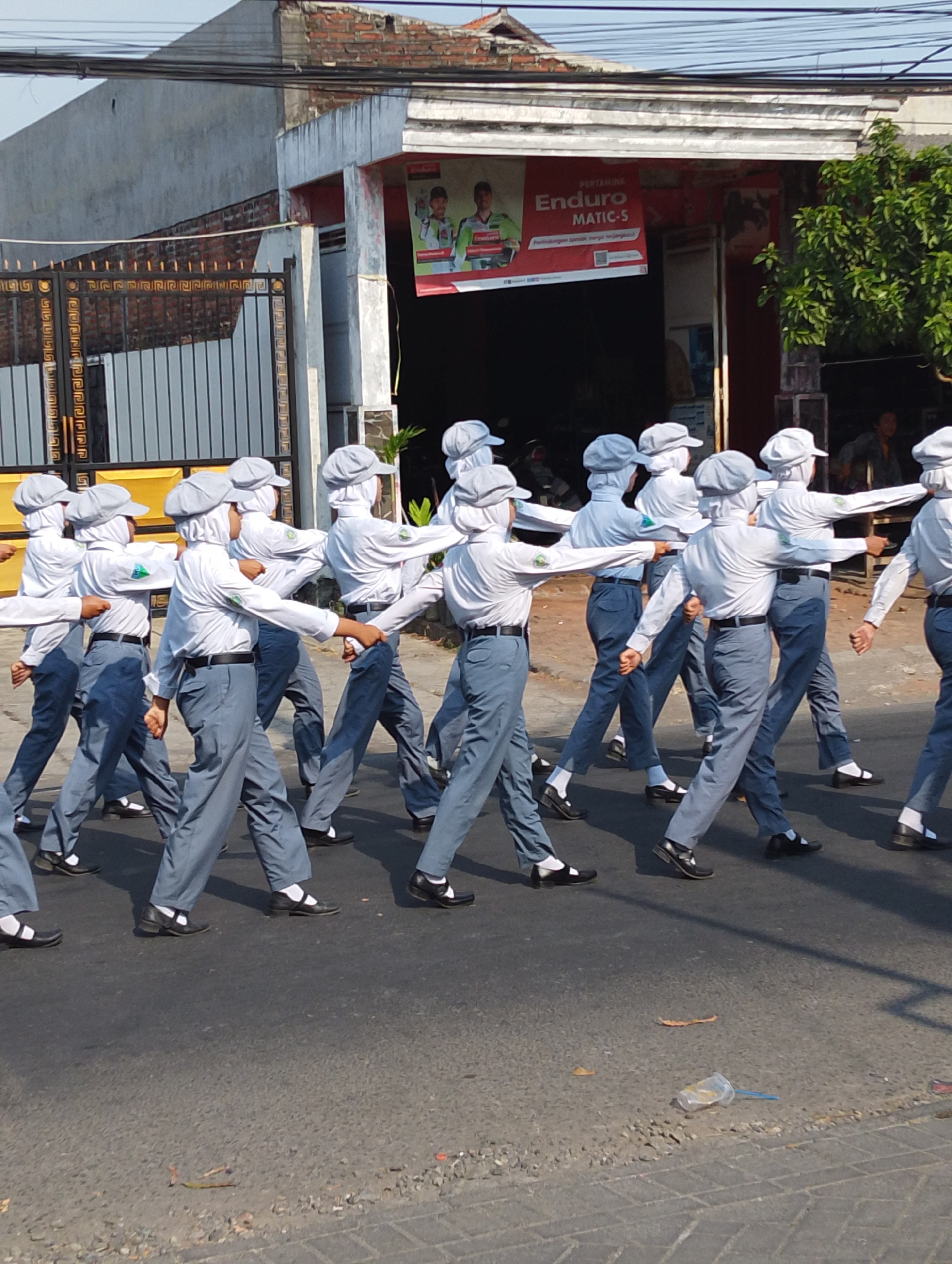 In Manaruwi, Indonesia, a group of uniformed students marches in formation on a paved street. They are wearing light-colored shirts, gray pants, and matching hats, with many of them holding their arms outstretched to the side. The students are walking in the direction of the viewer's right. In the background, there is a building with a red banner advertising "Enduro MATIC-S," and a large, ornate metal gate is visible to the left. The scene appears to be an outdoor event or parade.