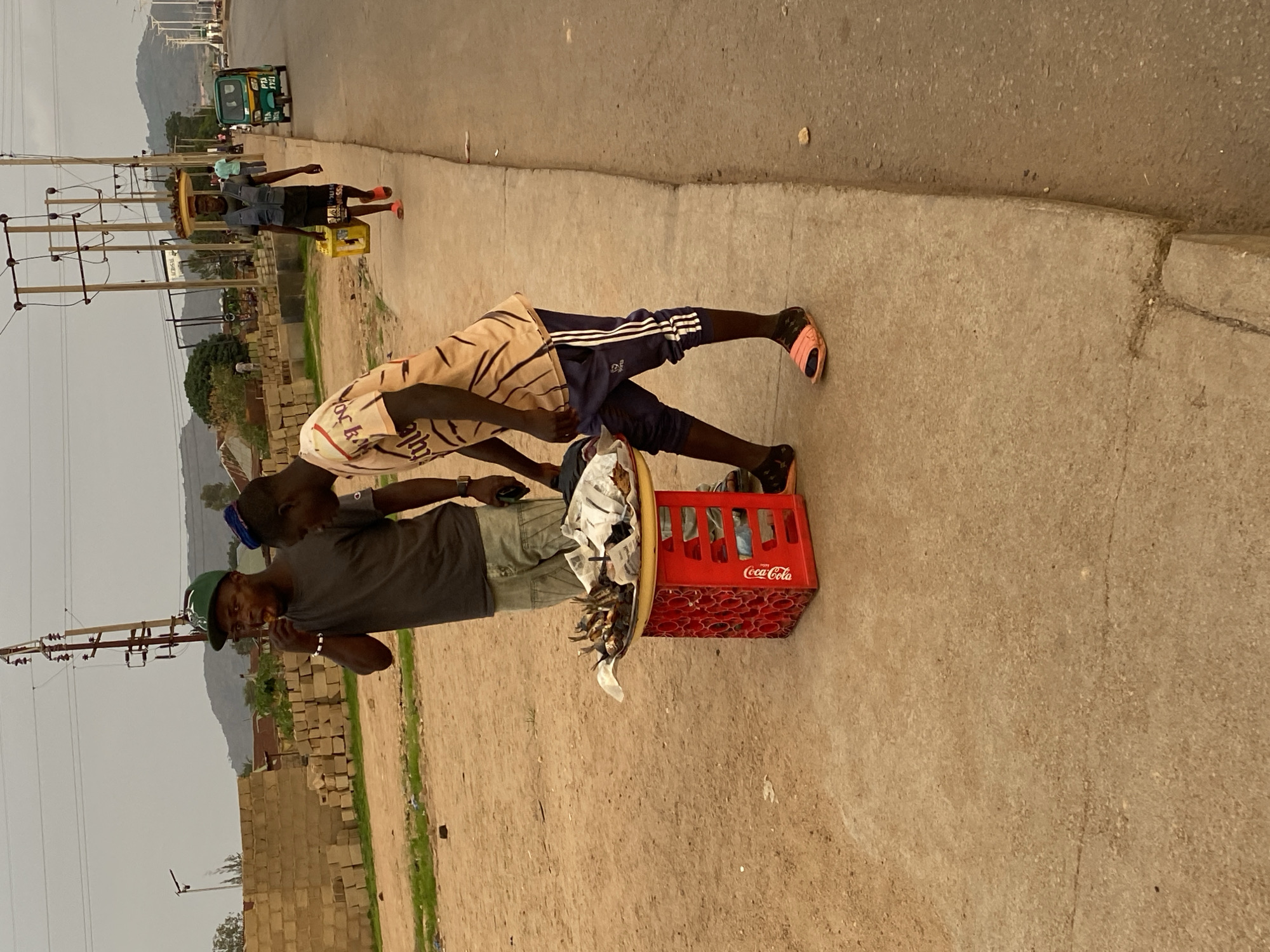The image captures a scene on a street in Bukuru, Nigeria. Two men stand next to a red Coca-Cola crate placed on the ground. One man, wearing a green hat and a gray shirt, appears to be holding something near his mouth. The second man, wearing an orange and white striped shirt and Adidas pants, has his hand on the first man’s shoulder. A tray filled with what looks like roasted or fried fish rests atop the crate. In the background, other individuals walk along the road near a yellow crate. A three-wheeled vehicle can be seen further down the road. Utility poles line the street, and piles of concrete blocks are visible on the side. The sky is overcast.