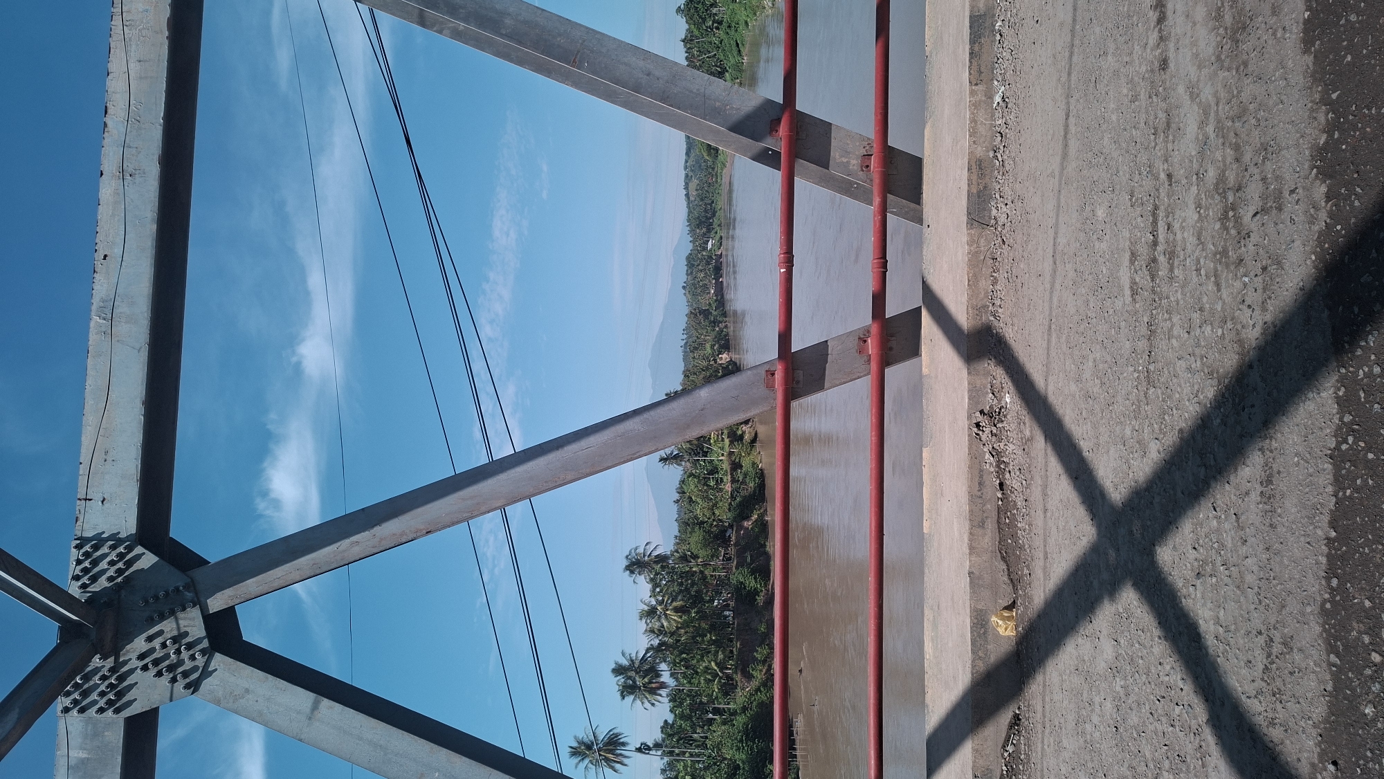 The image displays a view from a bridge overlooking a river in Kulukuta, Indonesia. The foreground features the bridge's steel structure, with large, weathered metal beams forming triangular supports. A dark, crisscrossed shadow from the bridge structure is cast upon the rough, gravelly surface of the bridge deck in the bottom portion of the frame. Red metal railings are visible just beyond the bridge deck.
The middle ground reveals a wide, muddy brown river flowing gently. The riverbanks are densely lined with lush green vegetation, primarily palm trees, characteristic of a tropical environment. In the distance, rolling hills or mountains are visible under a clear blue sky with scattered white clouds. A few power lines are strung across the sky, suggesting the presence of infrastructure. There are no people or vehicles visible in the scene. The overall impression is one of a serene, natural landscape dominated by the river and the surrounding greenery, viewed from an industrial structure.