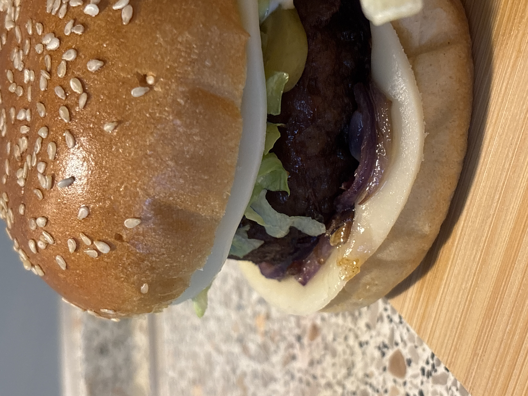 A close-up, eye-level shot shows a hamburger with a sesame seed bun. The hamburger is placed on a wooden board. The top bun is golden brown with many sesame seeds. The patty is dark brown, topped with lettuce, pickles, and onions. A slice of white cheese is visible under the patty. The background is blurred, showing a countertop. The image is taken in Wörth am Main, Germany.