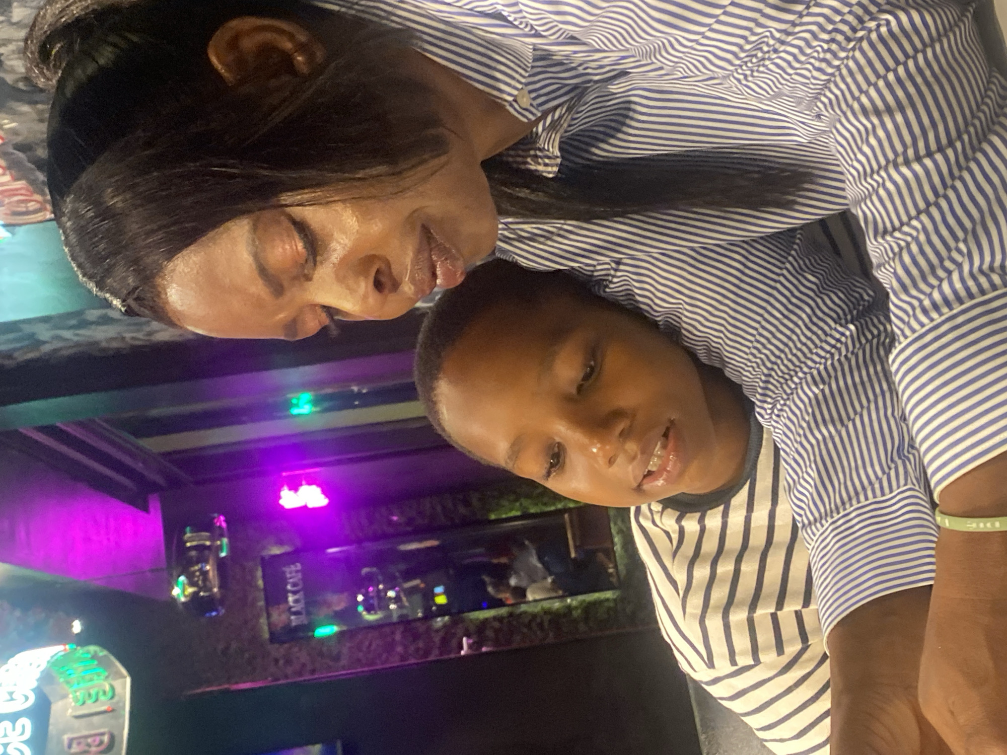 This image captures a close-up of a Black woman and a young Black boy, seemingly in a dimly lit indoor setting with vibrant neon lighting, possibly a restaurant or entertainment venue. The woman, positioned on the right, has dark hair pulled back and is wearing a blue and white striped shirt. Her head is tilted down, and she appears to be looking at something below the frame, her expression one of deep thought or concern. Her eye makeup is visible, with a shimmery orange-gold shadow.
The boy, on the left, is leaning against the woman, his head resting on her shoulder or chest. He is wearing a white t-shirt with thin dark stripes. His eyes are open, and he appears to be looking towards the woman or downwards. His mouth is slightly open, revealing some teeth.
In the background, a mirror reflects a scene with multiple people, some appearing to be seated. Neon signs are visible, one with green lettering that partially reads "ICE C..." and another that says "BLACK CAFÉ" in a frame. The overall atmosphere is dark, punctuated by the glow of purple and green lights, creating a moody ambiance. The presence of neon signs and the style of the interior decor suggest a modern urban environment. Ado, Nigeria, is noted as the location, hinting at a specific cultural context for this scene. The image is taken from a high angle, looking down slightly on the subjects.