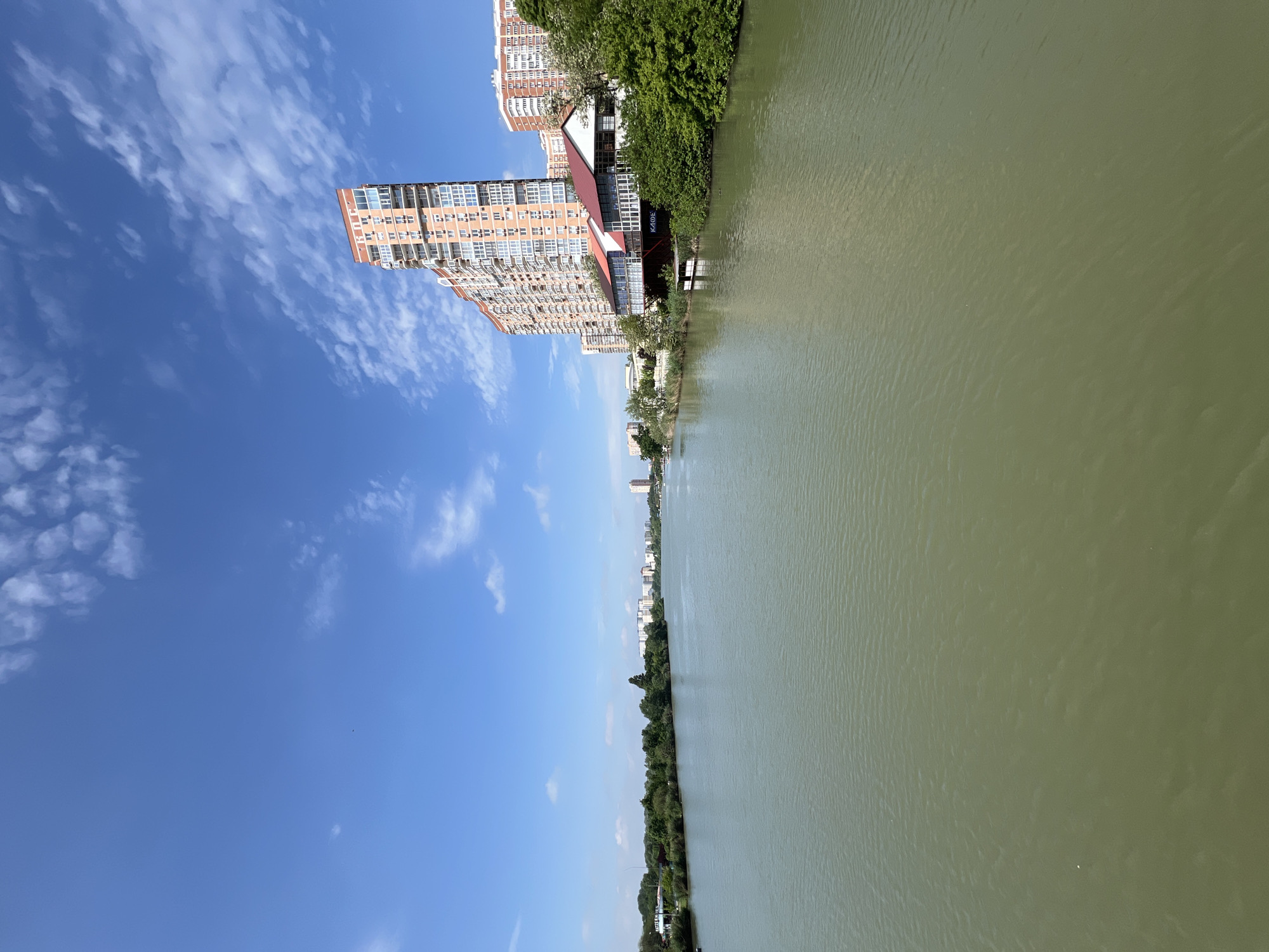 The image depicts a view of the Kuban River in Krasnodar, Russia. Along the riverbank, there is a mix of residential buildings, including high-rise apartments, and green vegetation. A building with a visible sign labeled "KAOE" (possibly a cafe) is also present. The sky is blue with scattered clouds. The river appears calm with a greenish hue. The image appears to be taken from a high vantage point.
