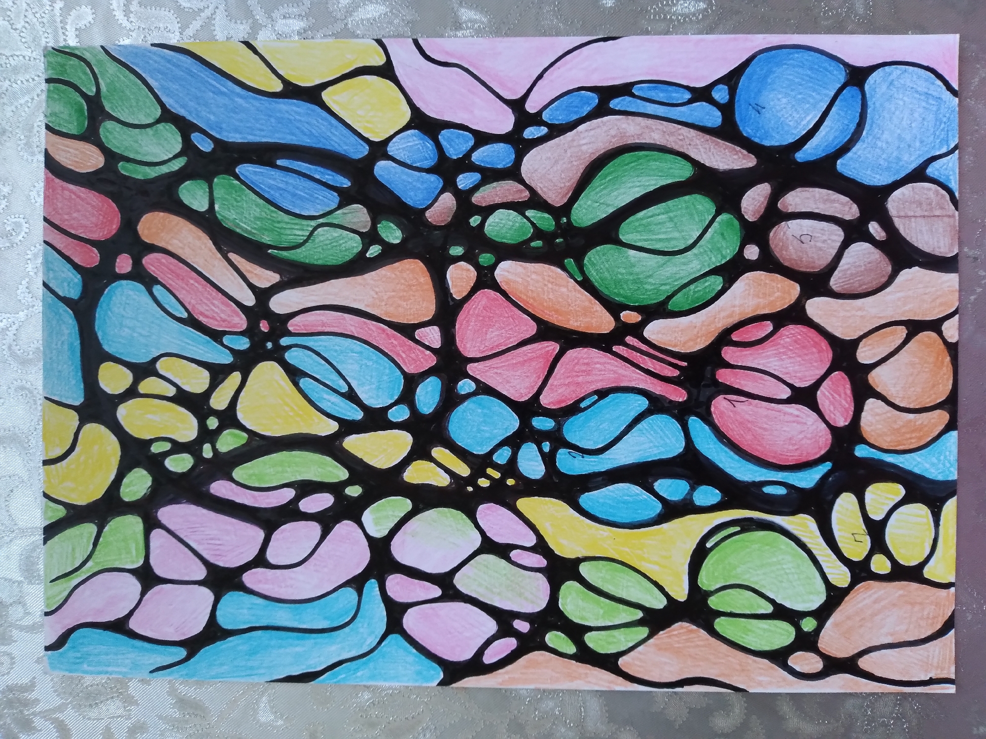 The image is a close-up, overhead shot of a piece of artwork on a textured, light-colored surface. The artwork is a drawing with black outlines creating an abstract pattern of connected, irregularly shaped cells. These cells are filled with various vibrant colors, including light blue, pink, yellow, green, orange, and a lighter blue. Some of the cells are outlined in black. The overall style is reminiscent of neurographic art or a stained-glass effect. The background surface appears to be a fabric with a subtle woven pattern. There are no people, actions, or interactions visible in the image. The artwork is the sole subject. There is no information or visual cues within the image that would indicate its location in the city of Baki, Uzbekistan.
