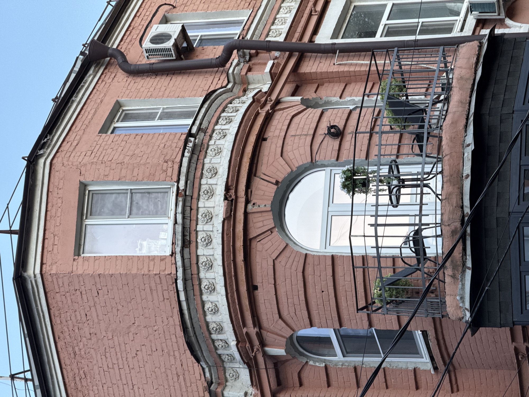This is a low-angle, medium shot of the corner of a historic pink brick building in Rostov-on-Don, Russia. The building features ornate architectural details, including a decorative band with floral motifs above a rounded balcony. The balcony, equipped with a black metal railing, has two black chairs, a small black table, and two potted plants. A large, arched window with a white frame is visible behind the balcony. Overhead, a brown downspout, an air conditioning unit, and several black electrical wires are present against a pale blue sky. The building's facade exhibits signs of wear, with some chipped paint and exposed brick.