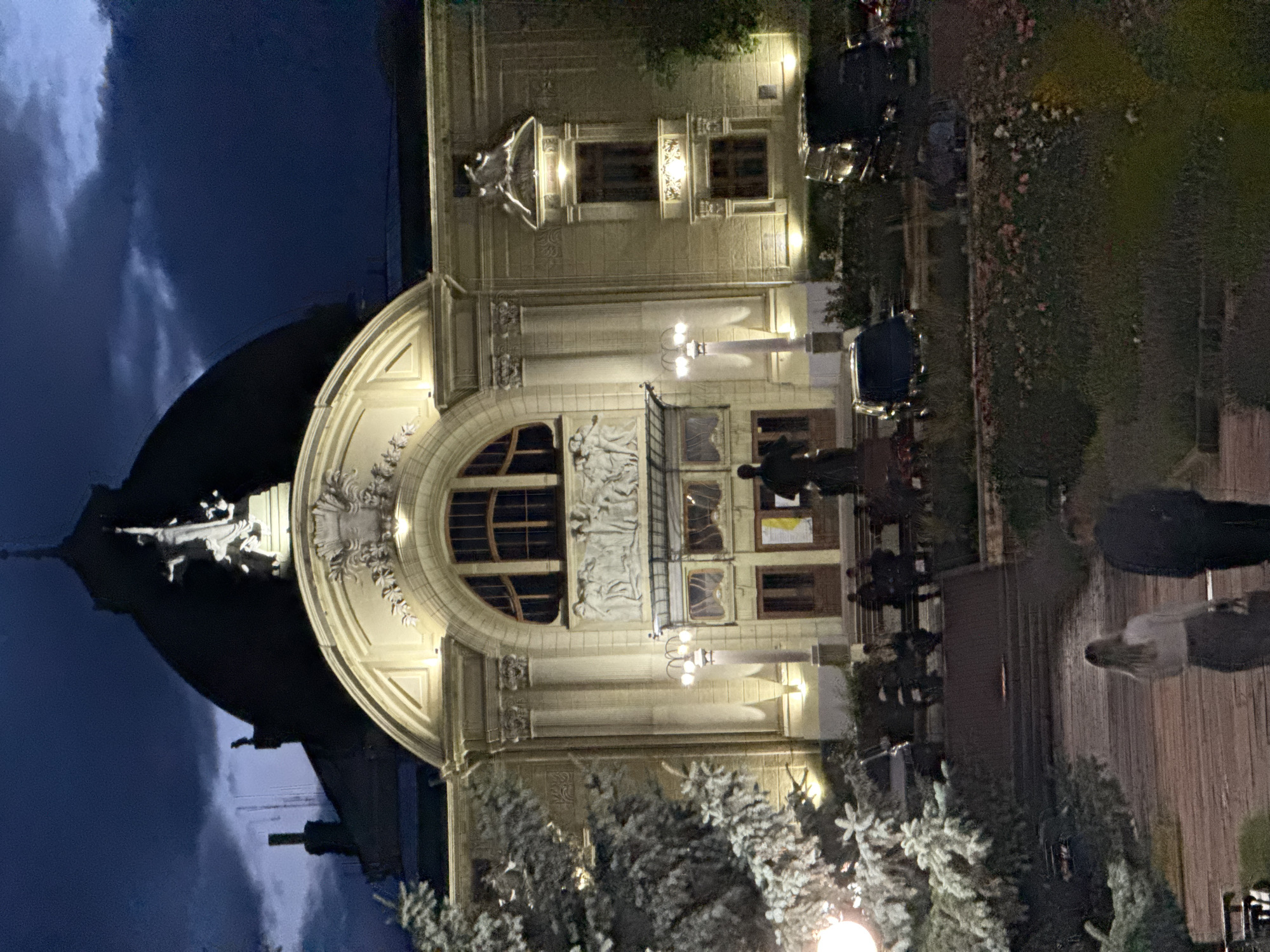 The image depicts a nighttime view of the Chernivtsi National University building, also known as the former residence of the Metropolitan of Bukovina and Dalmatia, in Chernivtsi, Ukraine. The building is illuminated by exterior lights, highlighting its ornate facade, large arched windows, and a prominent dome topped with a statue. In the foreground, a pathway leads towards the building, with two individuals walking away from the camera. A large evergreen tree is visible to the left of the frame. Several cars are parked in front of the building, and a few people are seated on benches near the entrance. The sky is dark with visible clouds, suggesting a twilight or evening setting.