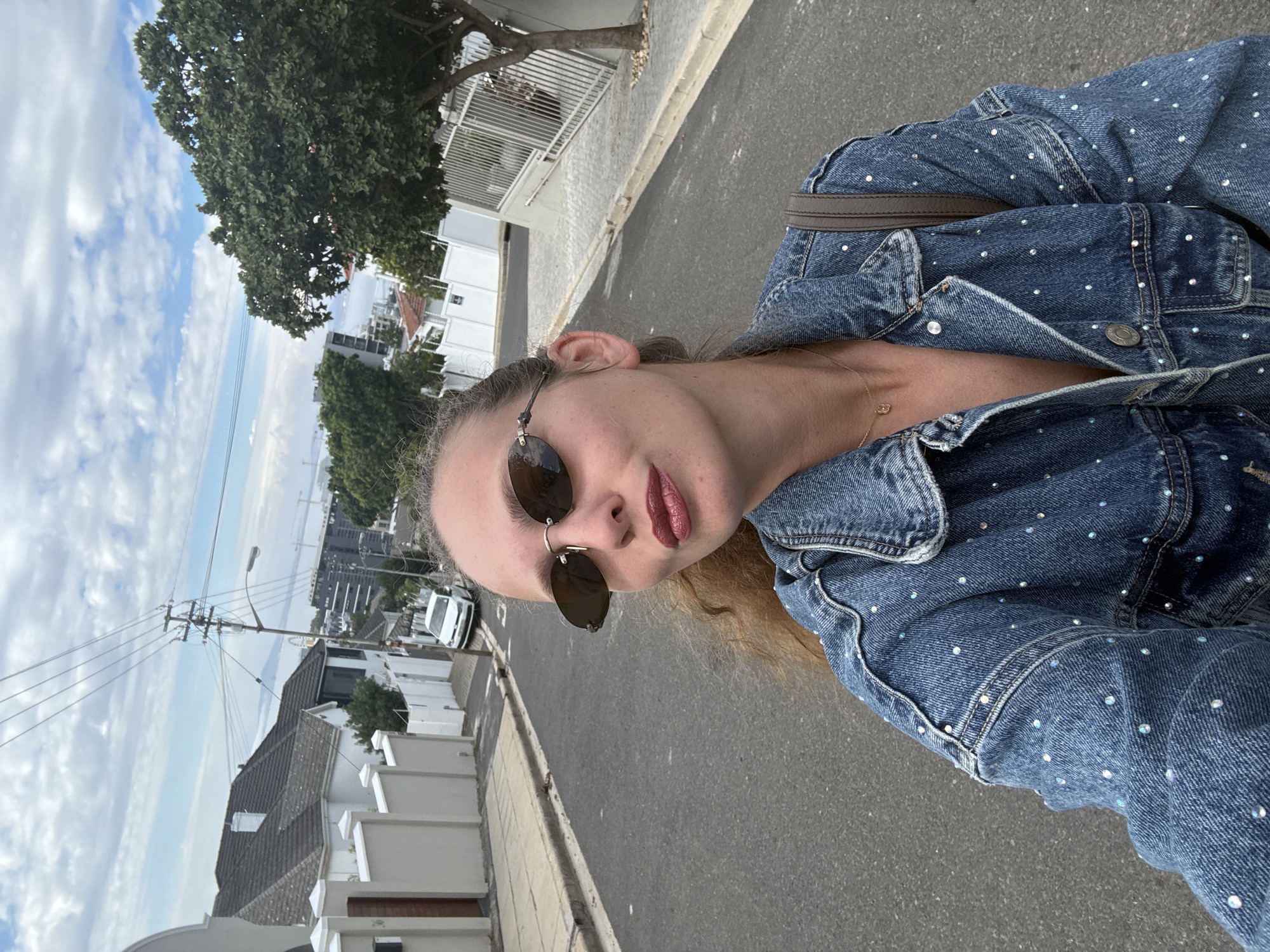 A medium-angle, candid outdoor shot depicts a person in a denim jacket adorned with rhinestones. The person is wearing sunglasses and appears to be standing on a street in a residential area. Buildings, trees, utility poles, and a cloudy sky are visible in the background, indicating an urban setting. The location is likely in Cape Town, South Africa, given the architectural style of the buildings.