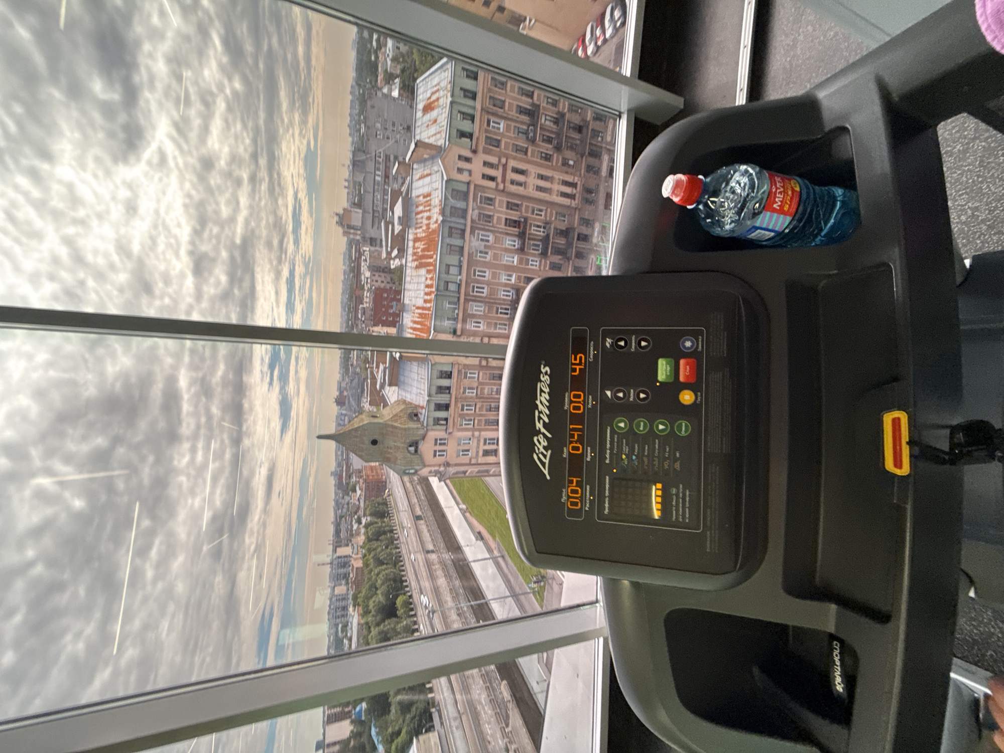 A close-up, eye-level shot shows the console of a Life Fitness treadmill in a gym. The treadmill display shows 0.04 miles, 0:41 time, and 4.5 speed. There is a bottle of water with a red cap in the cup holder on the right side of the console. The treadmill faces a large window that offers a panoramic view of a city. The sky is cloudy, and the city skyline includes various buildings, a highway overpass, and trees. The overall impression is a modern gym with an urban view.