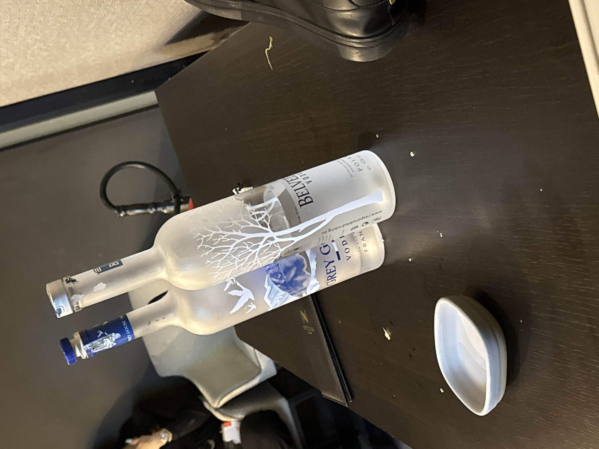 Two bottles of vodka, one Grey Goose and one Belvedere, are placed on a dark wooden table. A small, light-colored dish sits in the foreground, and a black shoe is visible on the right side of the image. In the background, a hookah hose is visible. The scene appears to be indoors, possibly in a lounge or bar. There is no clear indication of the location in Sofia, Bulgaria.