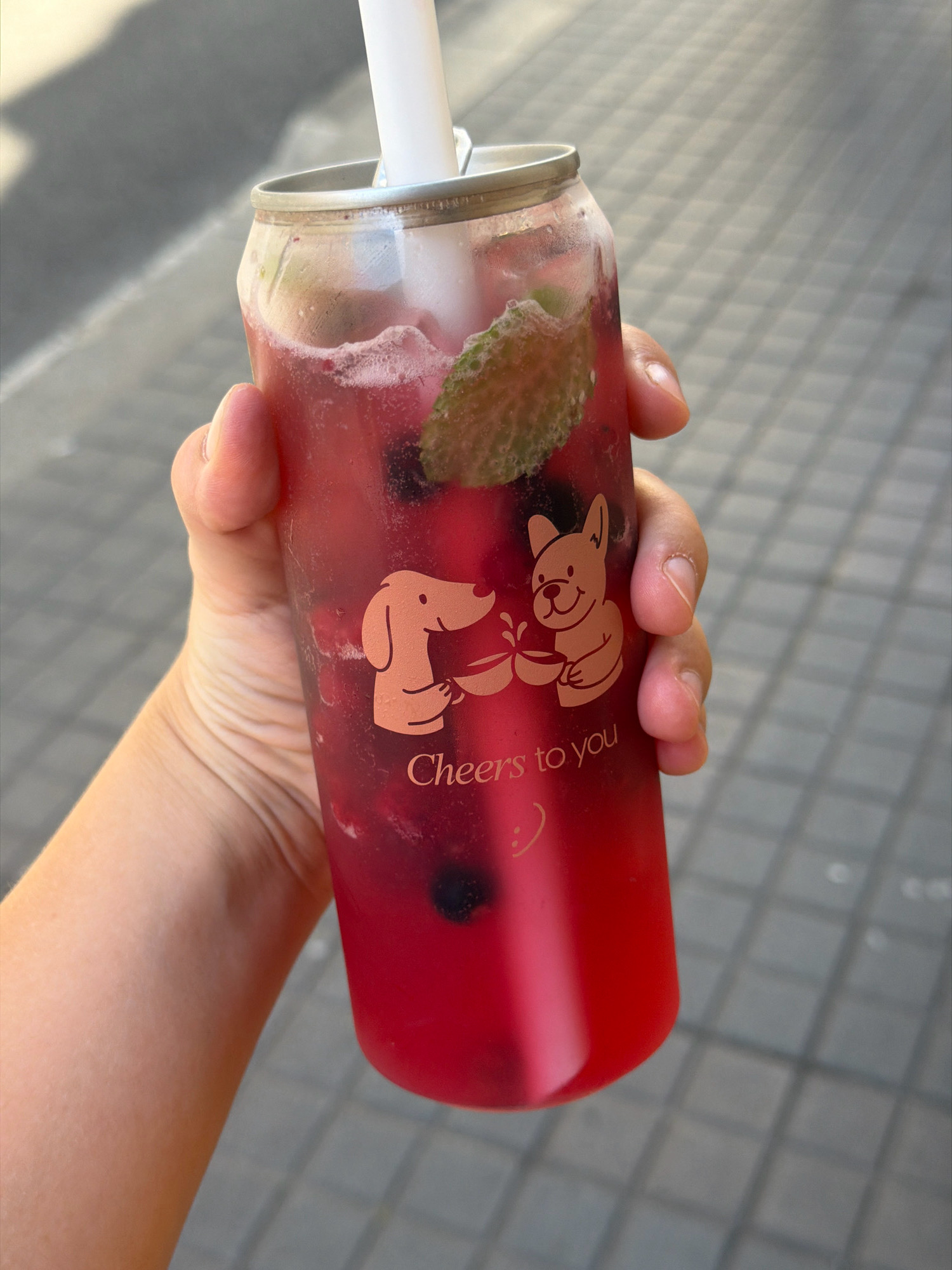 A person's hand holds a translucent, can-shaped cup filled with a vibrant red beverage, appearing to be a mixed drink with berries and mint. The cup has an imprint of two cartoon dogs and the text "Cheers to you" with a smiley face. A white straw extends from the top of the cup. The background consists of a paved surface with a checkered pattern, suggesting an outdoor urban setting, possibly a sidewalk in Barcelona, Spain. The lighting indicates a bright, daytime scene.