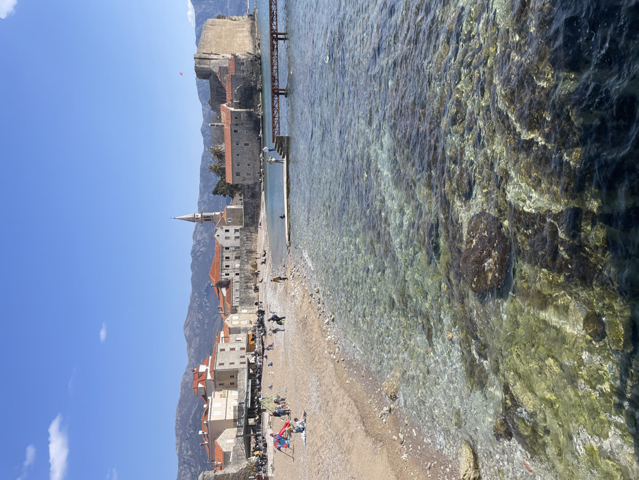 The image displays a sunny coastal scene in Budva, Montenegro, featuring the Old Town (Stari Grad) from a beach perspective.
**Environment:**
The sky is clear blue with sparse, wispy white clouds. Distant mountains with visible peaks frame the background. The Old Town, characterized by stone buildings with orange-tiled roofs, a prominent church steeple on the left, and fortified walls, lines the coastline. A large stone fortress, identified as the Budva Citadel, is situated at the easternmost point of the Old Town, extending into the sea. A light-colored sand and pebble beach, Ricardova Glava, stretches along the Old Town's fortifications. The foreground features clear, shallow, turquoise-to-green ocean water, revealing submerged rocks and dark patches of aquatic vegetation. The water darkens with increasing depth further from the shore.
**Objects and Structures:**
A wooden pier with a concrete section extends into the water from the right side of the beach. A smaller, L-shaped concrete pier is visible closer to the shore. A colorful playground structure (red, blue, yellow) is present on the sand in the mid-left. A large, dark-colored awning covers an outdoor seating area on the beach, adjacent to the Old Town wall.
**People and Actions:**
Numerous individuals are dispersed across the beach. Children are present near the playground structure and along the water's edge. Adults are seen walking, sitting, and congregating under the awning, suggesting dining or socializing. On the longer pier, at least two individuals are standing on the concrete section, while one person is seated on the smaller concrete pier. The overall scene indicates recreational activity on a bright day.