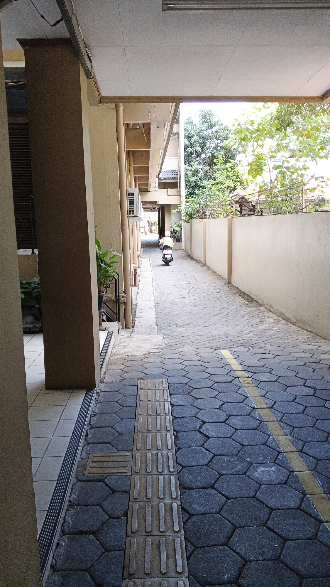 The image shows a narrow paved walkway or alley between two buildings. A person is riding a white scooter away from the camera down the alley. The walkway is paved with hexagonal dark grey bricks and a yellow line runs along the right side of the walkway. There is a tactile paving strip on the left side of the walkway. The buildings on either side are light beige in color. The building on the left has a large beige pillar in the foreground and an air conditioning unit visible. The building on the right has a low beige wall. Trees and foliage are visible beyond the wall on the right. The lighting suggests it is daytime. The scene appears to be in a residential or commercial area, possibly a back alley or a pedestrian walkway within a complex in the city of Yogyakarta, Indonesia.