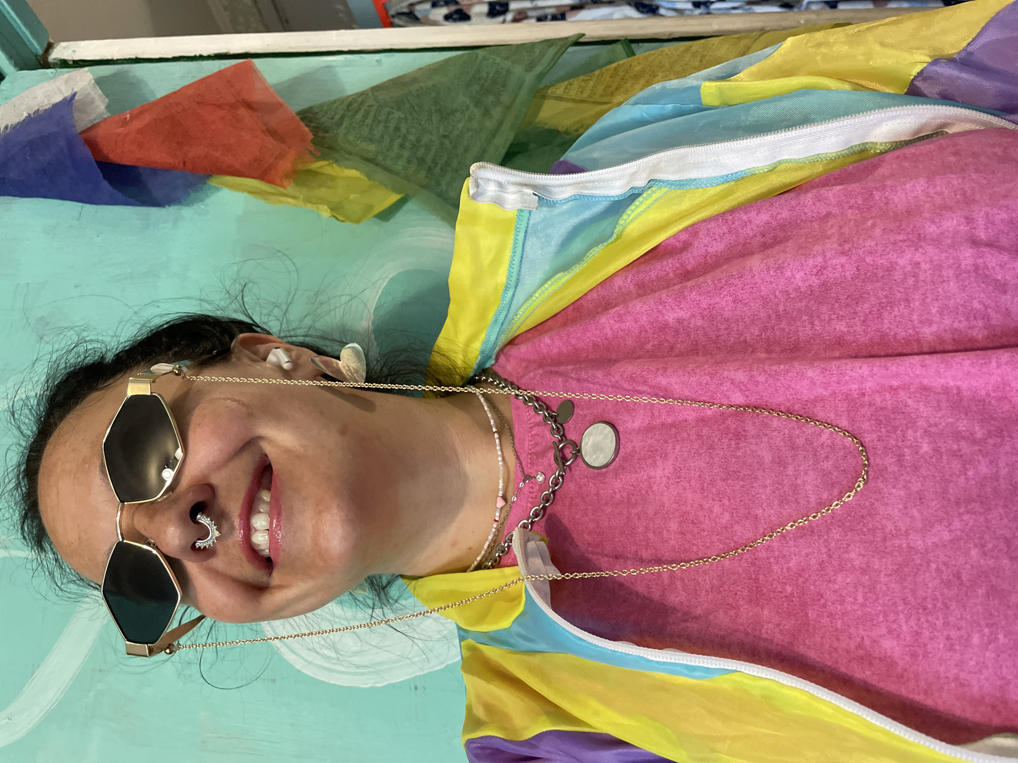 A woman with dark hair is pictured from the chest up, wearing an open, colorful windbreaker over a bright pink t-shirt. She has on polygonal-shaped sunglasses with gold frames and a septum piercing. The background consists of a teal-colored wall with white abstract patterns and several prayer flags in red, blue, yellow, and green. The woman is smiling and looking towards the viewer. The scene appears to be indoors, likely in a home or studio. There is no identifiable information to suggest this location is in Saint Petersburg, Russia.
