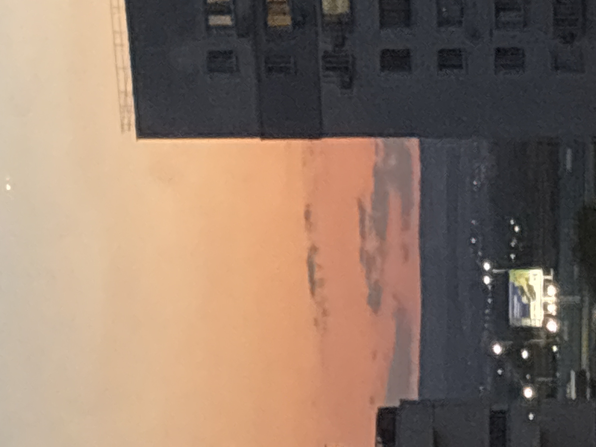 The image depicts a cityscape at dusk, with a prominent apartment building on the right and another building partially visible on the left. The sky exhibits a gradient of orange and pink hues, indicative of a sunset. In the mid-ground, a billboard is illuminated, surrounded by streetlights, suggesting an urban environment. The background shows a silhouetted landscape, possibly hills or a distant city. The overall impression is that of a residential area within a city. The image does not contain any people or specific actions.
