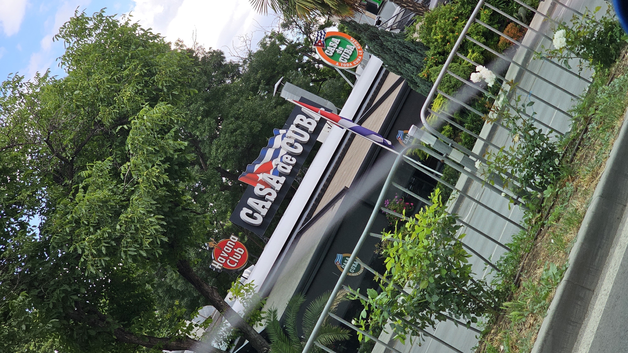 This is a photograph of the exterior of a establishment called "CASA de CUBA". The sign for the establishment is large and prominent, featuring stylized lettering and flags. Below the main sign, a smaller circular sign reads "Havana Club". The building appears to be a restaurant or bar, with a dark facade and windows. Green trees and foliage surround the building, suggesting an outdoor setting. A metal fence runs in front of the building, separating it from a street or pathway. White roses are visible growing along the fence. The sky is partly cloudy. The overall impression is that of a commercial establishment with a Cuban theme. The text "CASA de CUBA", "Havana Club", "CASA CUBA", and "TODA CUBA LE GUSTA" are visible.