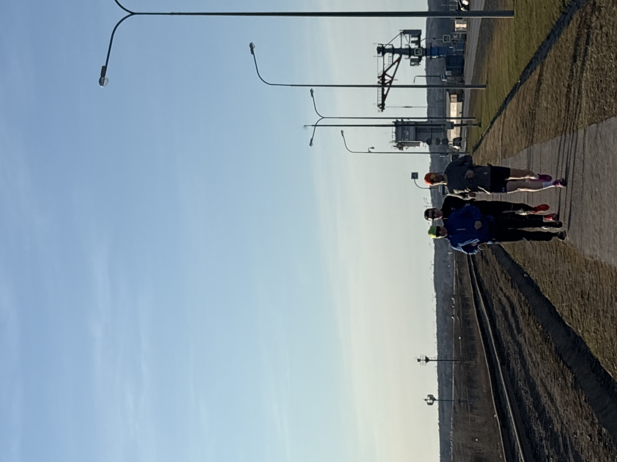 The image shows three runners on a paved path alongside a railway track. The location is Chaikovsky, Russia, identifiable by the industrial structures and the presence of a railway line. The sky is blue and clear. The runners are wearing sportswear and running shoes. They are running in the same direction. The path runs parallel to a grassy embankment. Street lights are visible along the path. Industrial structures are visible in the distance beyond the embankment.